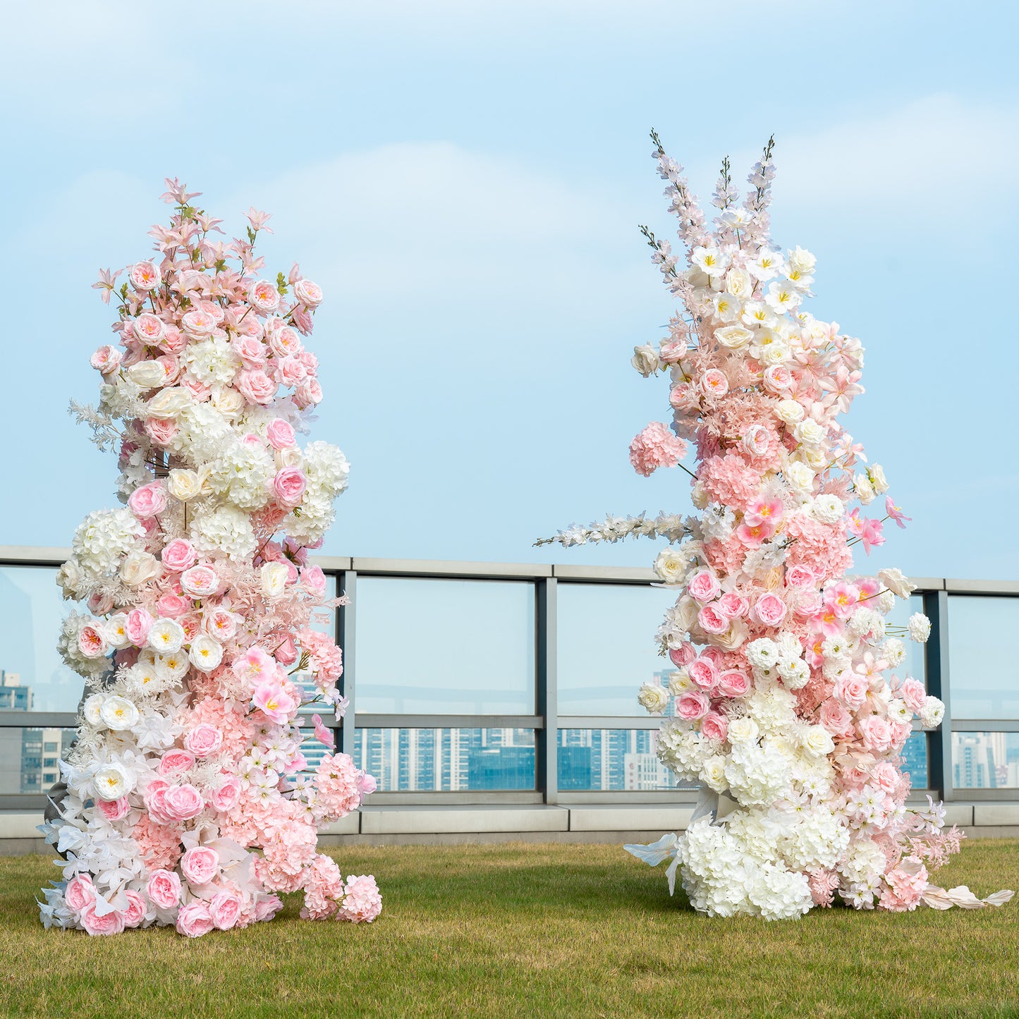 This backdrop features two tall floral arrangements placed outdoors on a rooftop with a cityscape in the background. The arrangements are composed of artificial flowers, including pink and white roses, white hydrangeas, and delicate pink blossoms that might be cherry - like flowers. The color palette is soft, with various shades of pink and white, creating a romantic and elegant style, perfect for wedding ceremonies or engagement photo shoots. 