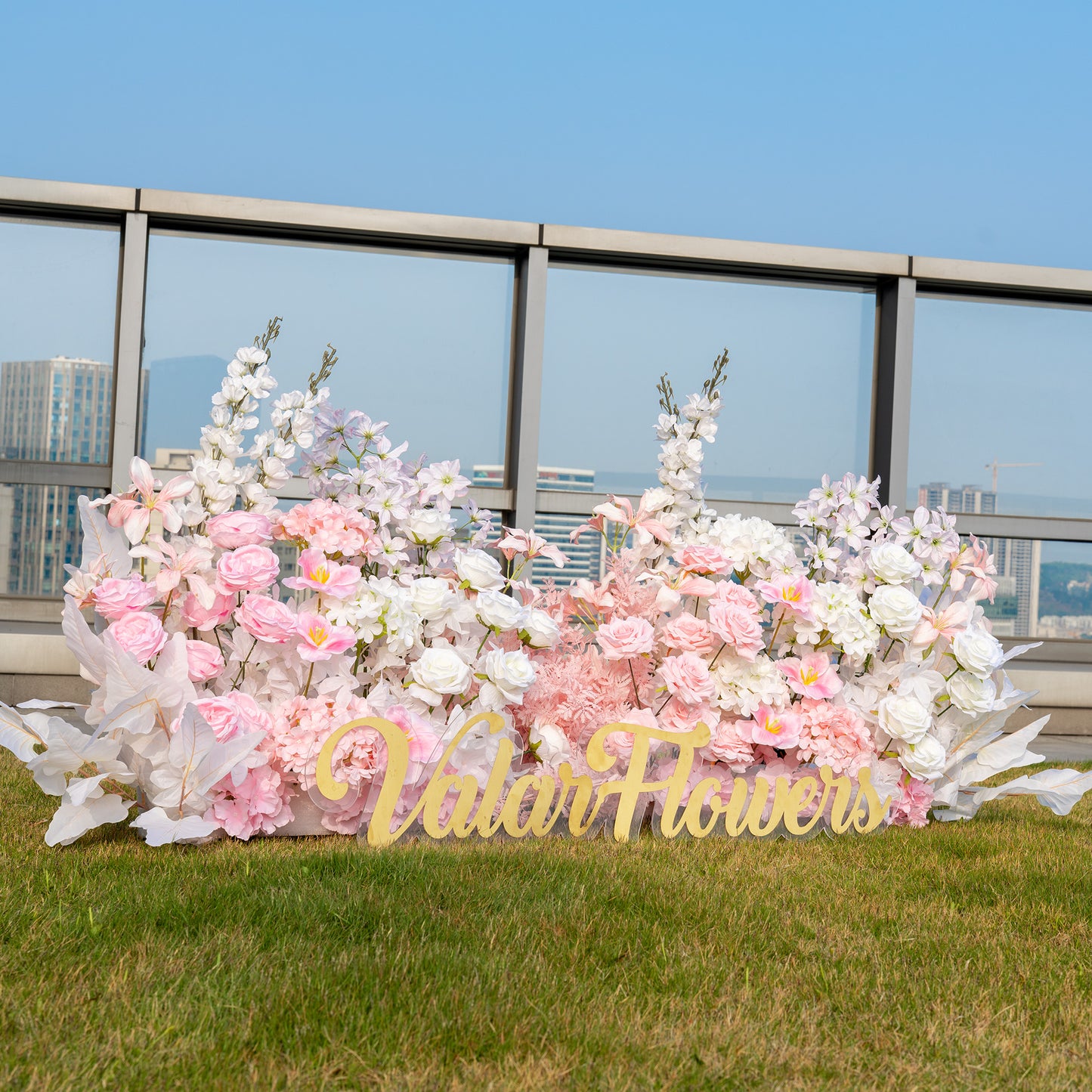 This backdrop, set on a rooftop with a cityscape in the background, features a lush arrangement of artificial flowers. It includes pink and white roses, white hydrangeas, and delicate pink blossoms. The color palette is soft, with pastel - toned pinks and whites, creating a romantic and elegant style. The “ValarFlowers” sign adds a branding touch. 