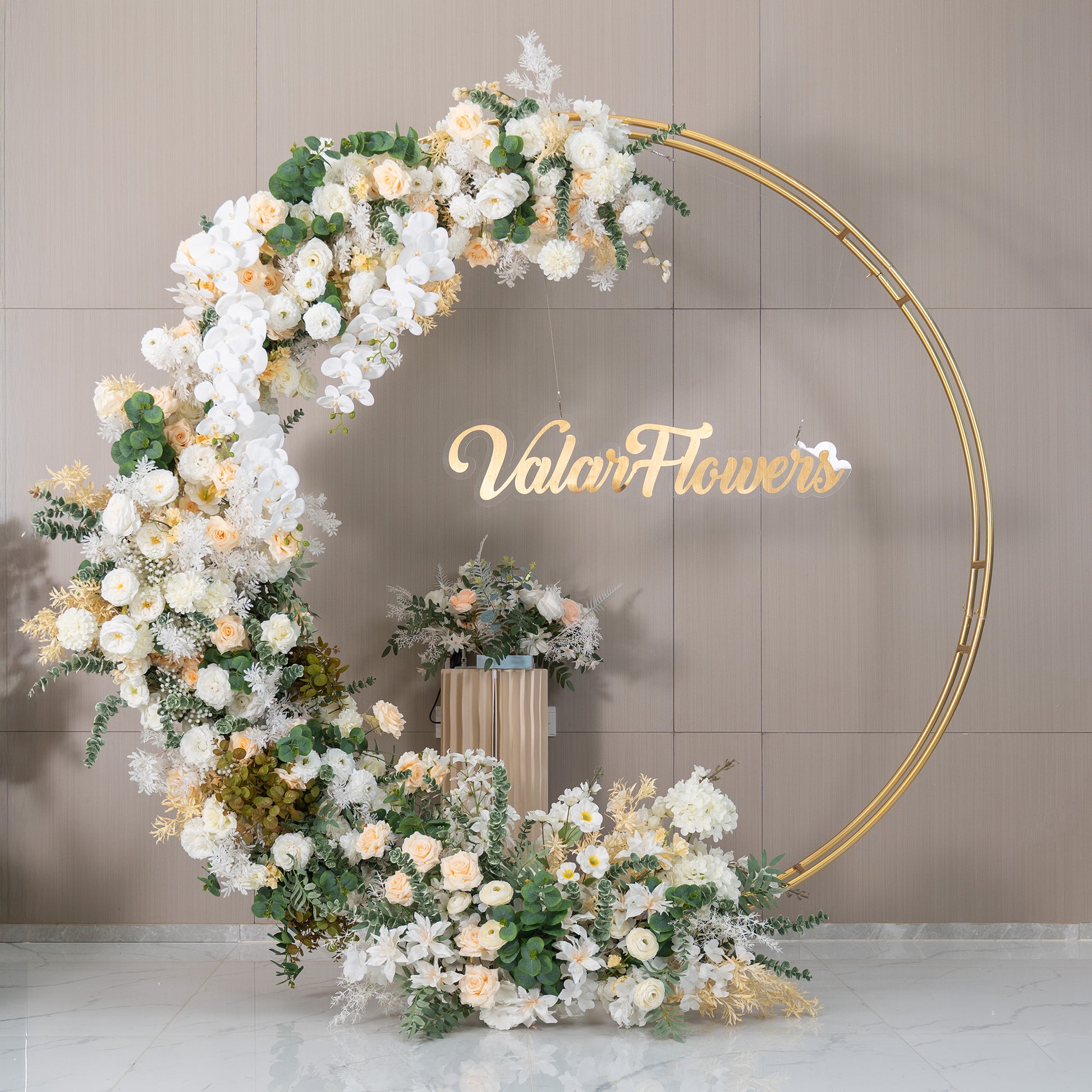 This backdrop features a golden - circular frame adorned with a lush arrangement of artificial flowers. Prominent flowers include white orchids, soft - hued roses in ivory and light peach, and white chrysanthemums. Interspersed with green eucalyptus and other foliage, the color palette is soft and elegant. The "ValarFlowers" sign adds a branded touch. The style is romantic and sophisticated, ideal for weddings or upscale events. 