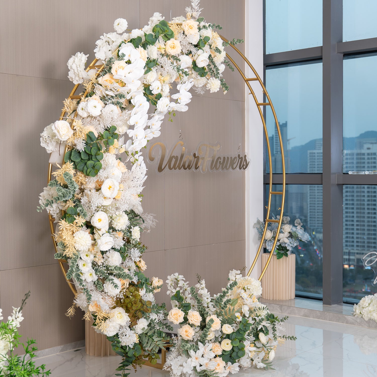 This backdrop features a golden circular frame adorned with an abundance of artificial flowers. Prominent flowers include white orchids, creamy - white roses, and white chrysanthemums. The color palette is dominated by soft whites, ivories, and hints of green from the foliage. The &quot;ValarFlowers&quot; sign adds a branded touch. The style is elegant and romantic, suitable for events like weddings. 
