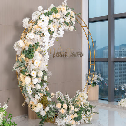 This backdrop features a golden circular frame adorned with an abundance of artificial flowers. Prominent flowers include white orchids, creamy - white roses, and white chrysanthemums. The color palette is dominated by soft whites, ivories, and hints of green from the foliage. The &quot;ValarFlowers&quot; sign adds a branded touch. The style is elegant and romantic, suitable for events like weddings. 