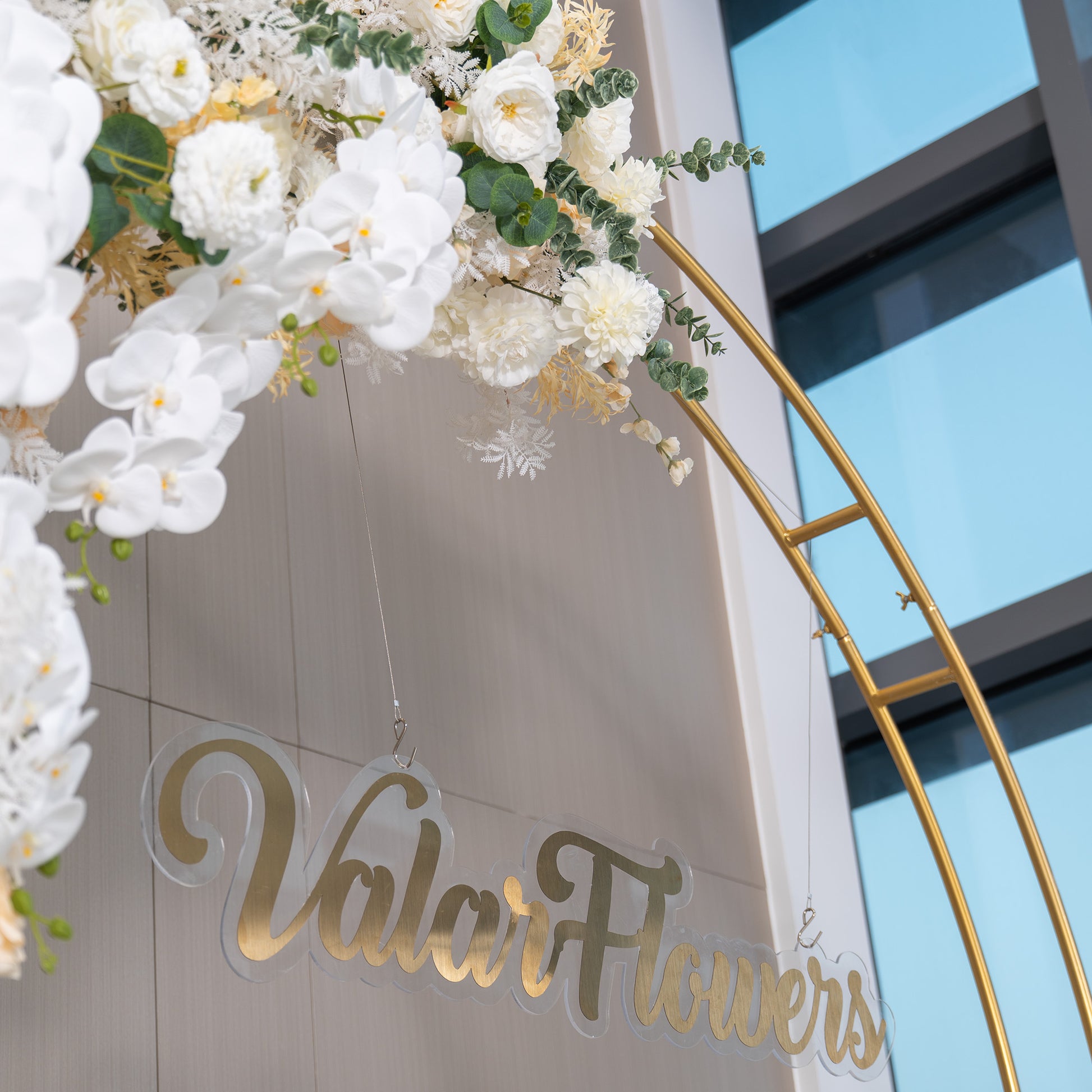 This backdrop features a golden - arched structure adorned with a lush arrangement of artificial flowers. Predominant are white orchids, complemented by white roses and chrysanthemums. Green eucalyptus leaves add a touch of natural greenery. The "ValarFlowers" sign in gold script hangs below. The style is elegant and sophisticated, with a soft, monochromatic color scheme. 
