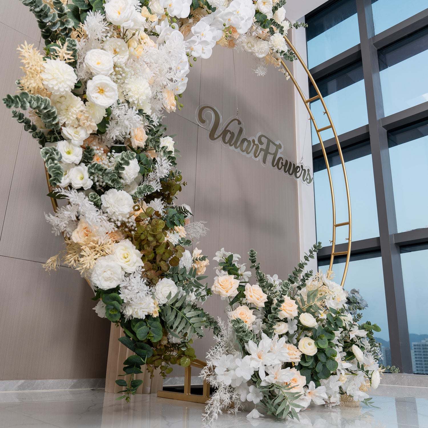 This backdrop features a golden - hued circular frame forming a crescent - like shape, adorned with an abundance of artificial flowers. Prominent flowers include white roses, white orchids, and pale - yellow roses, complemented by green eucalyptus and white fern - like foliage. The color palette is soft, with whites, ivories, and greens. The style is elegant and romantic, perfect for upscale events. 