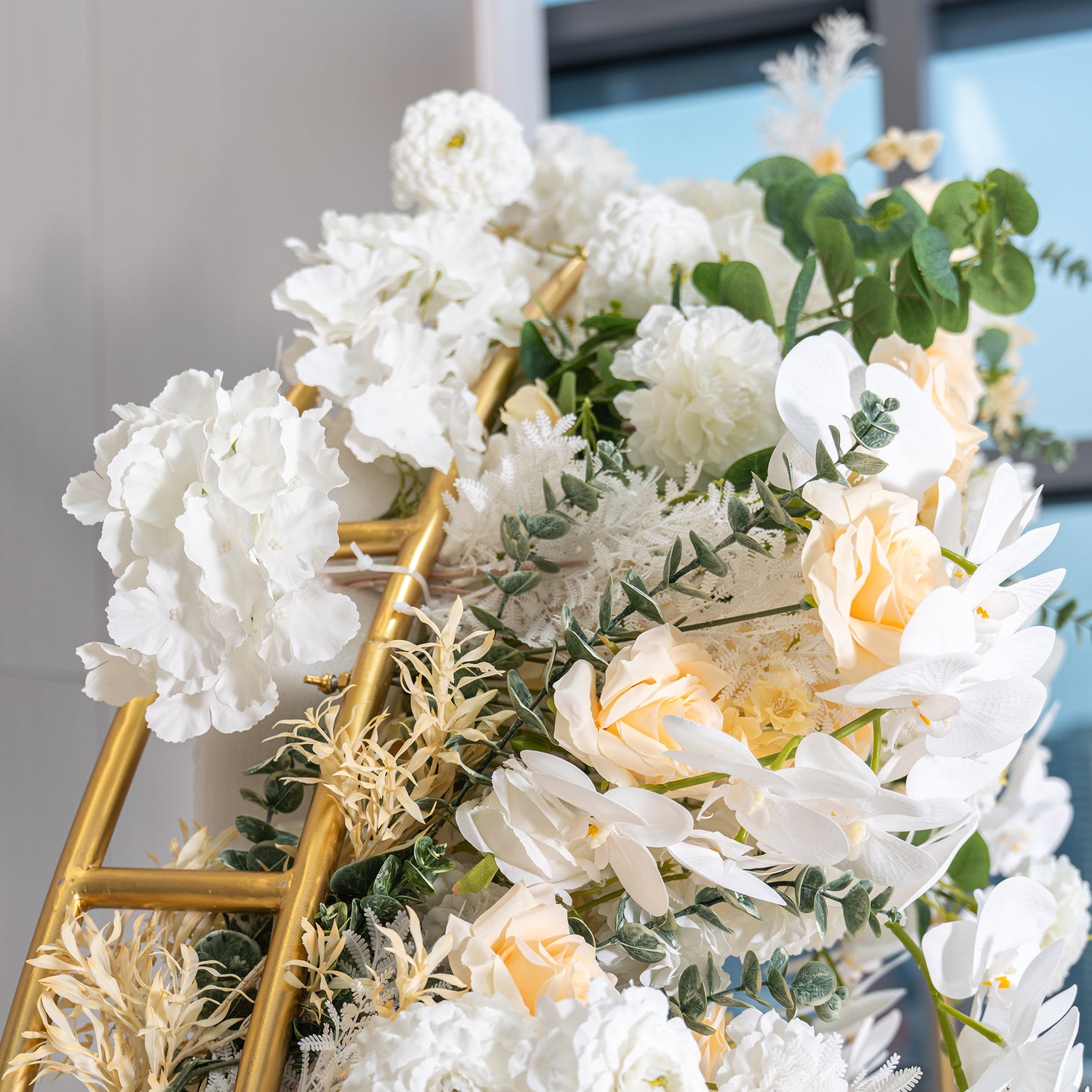 This backdrop features a golden - framed structure adorned with an array of artificial flowers. Prominent are white orchids, creamy - hued roses, and white hydrangeas. Interspersed are green eucalyptus leaves and delicate white fern - like foliage. The color palette is soft, with whites, ivories, and hints of green. The style is elegant and refined, perfect for upscale events. 