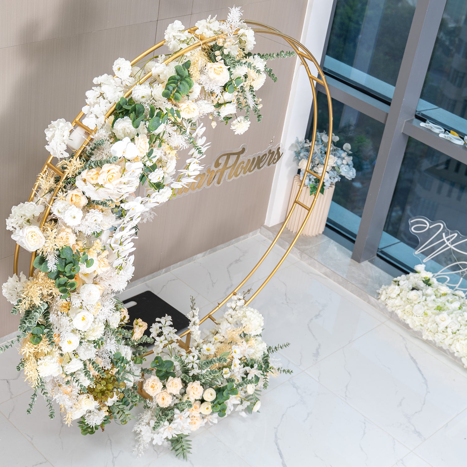 This backdrop features a golden - hued circular metal frame shaped like a crescent moon, adorned with an abundance of artificial flowers. The flowers include white roses, white orchids, and pale - yellow roses, complemented by green eucalyptus and white - tinged foliage. The color palette is soft and neutral, with whites, ivories, and greens. The style is elegant and romantic, ideal for weddings or upscale events. 