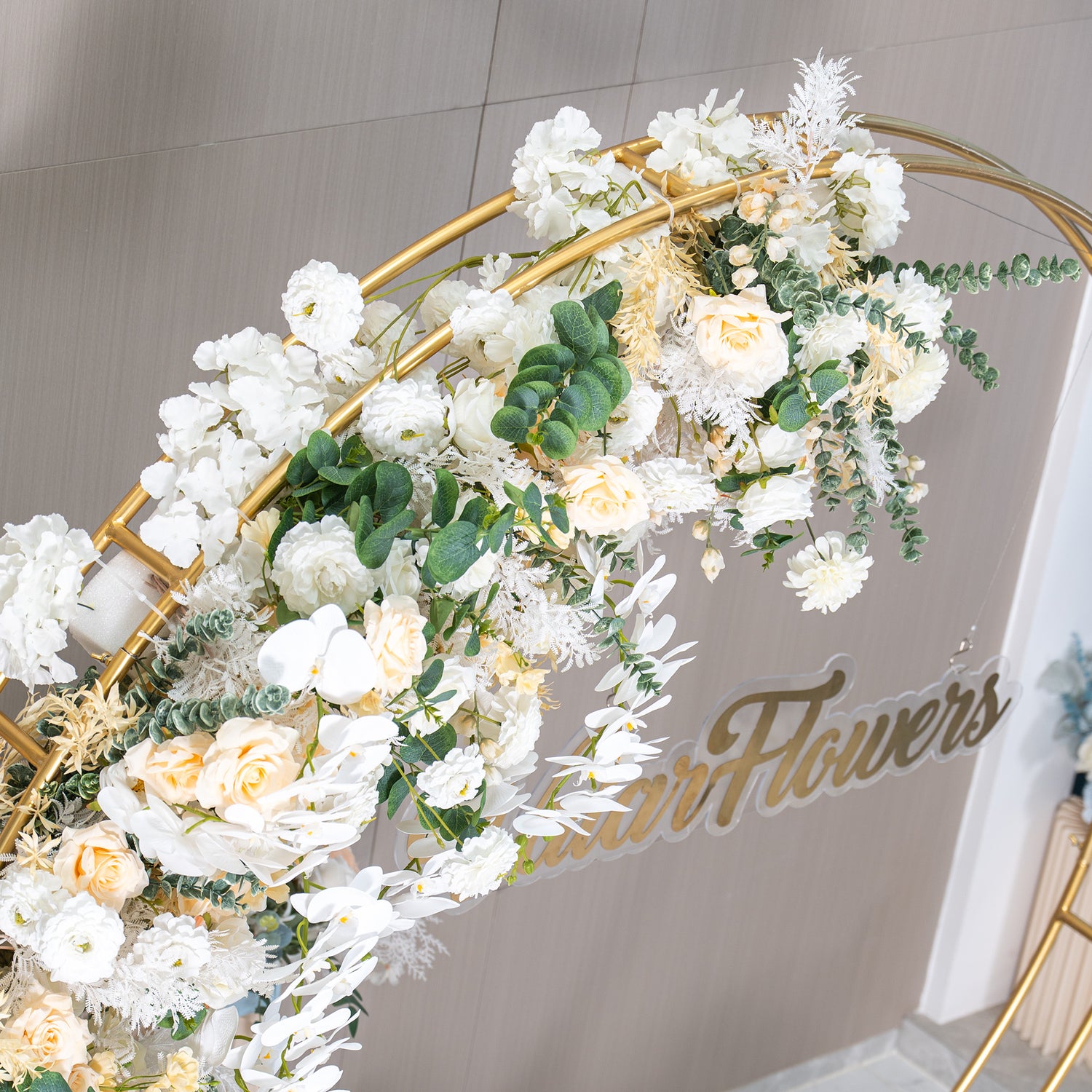 This backdrop features a golden - hued circular metal frame partially adorned with a lush arrangement of artificial flowers. Prominent flowers include white orchids, creamy - white roses, and white chrysanthemums. Green eucalyptus and other foliage add a touch of nature. The color palette is soft, with whites, ivories, and greens. The style is elegant and romantic, ideal for events like weddings. 