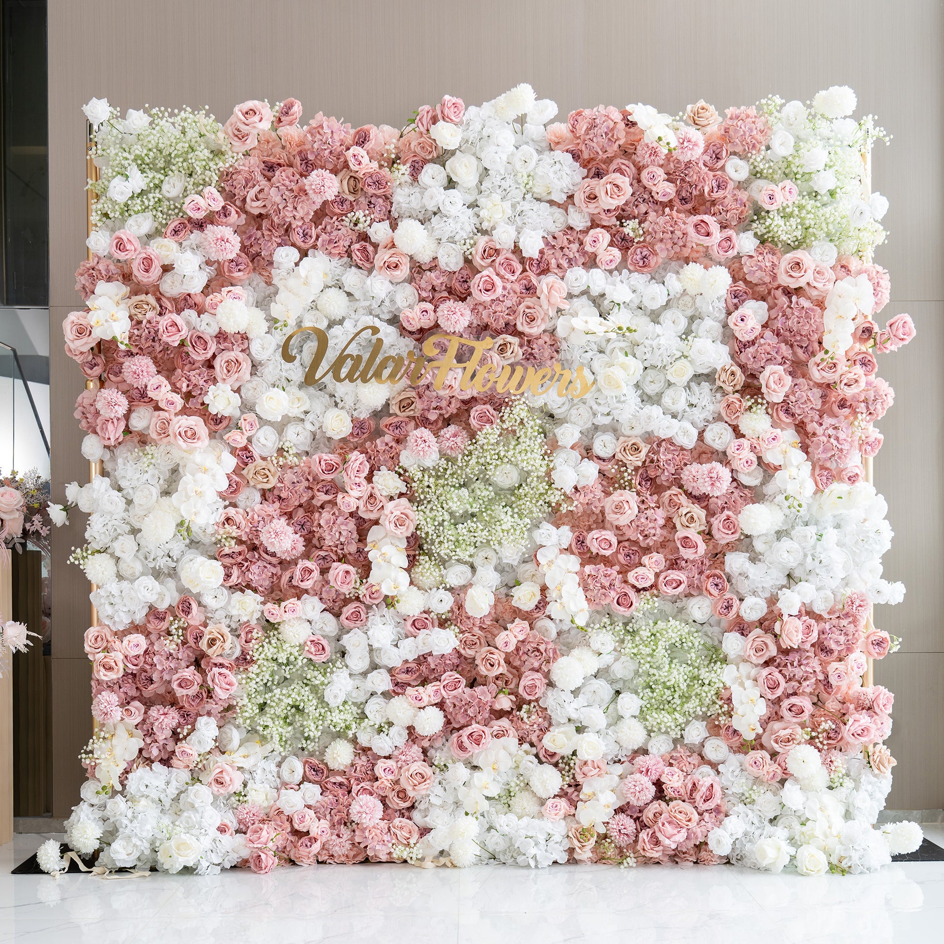 This backdrop is a square - shaped floral wall densely packed with artificial flowers. It features roses in various shades of pink, along with white roses and hydrangeas. Small clusters of baby's breath add a delicate touch. The color palette is soft, with pinks, whites, and hints of green. The style is romantic and charming, ideal for events like weddings or parties. The "ValarFlowers" sign in gold adds a branded element. 