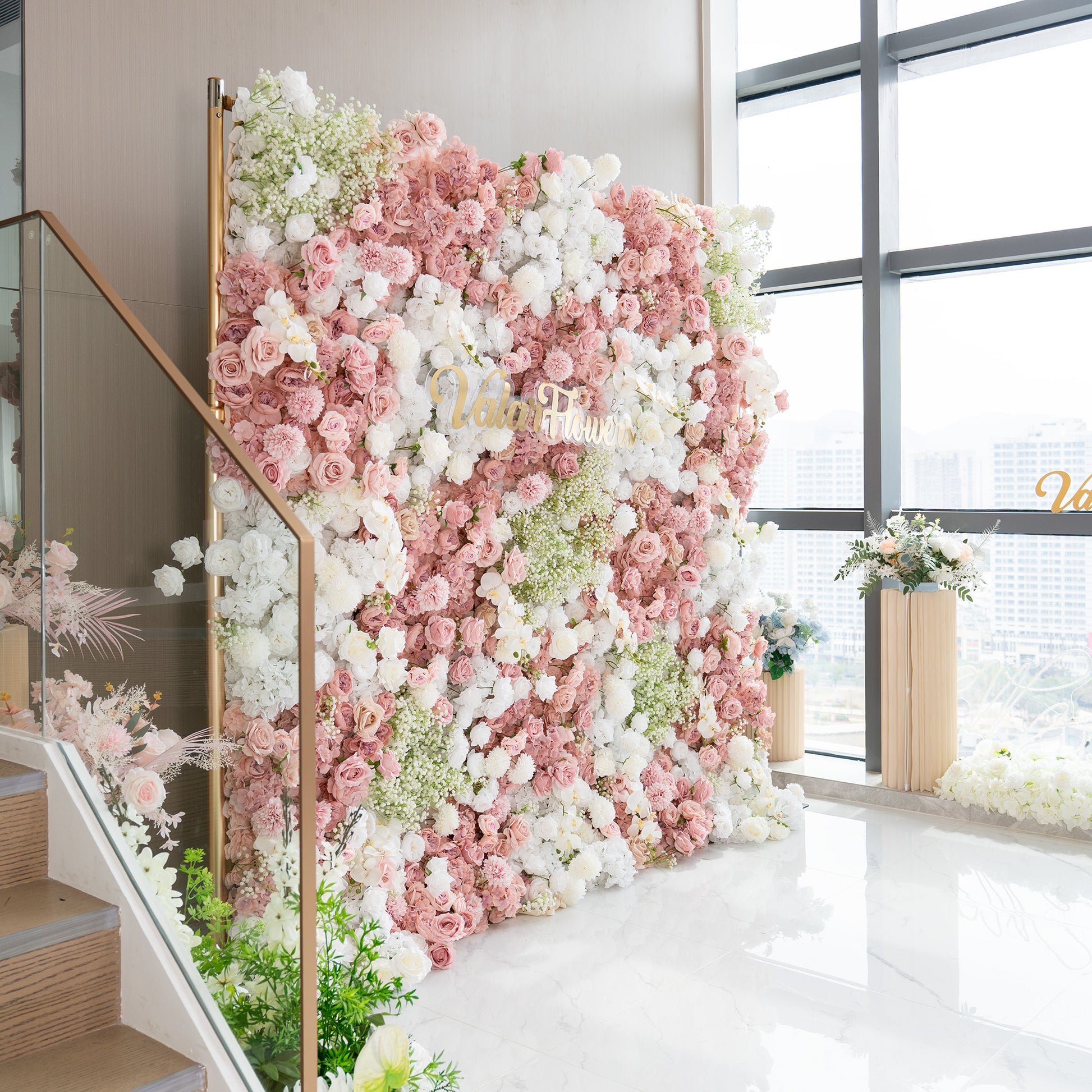This backdrop is a vertical floral wall supported by a metallic gold frame. It is densely adorned with artificial flowers, including roses in various shades of pink and white, along with clusters of baby's breath. The color palette features soft pinks, pure whites, and touches of green. The style is romantic and elegant, perfect for weddings or special events. The "ValarFlowers"  sign in gold adds a branded touch. 