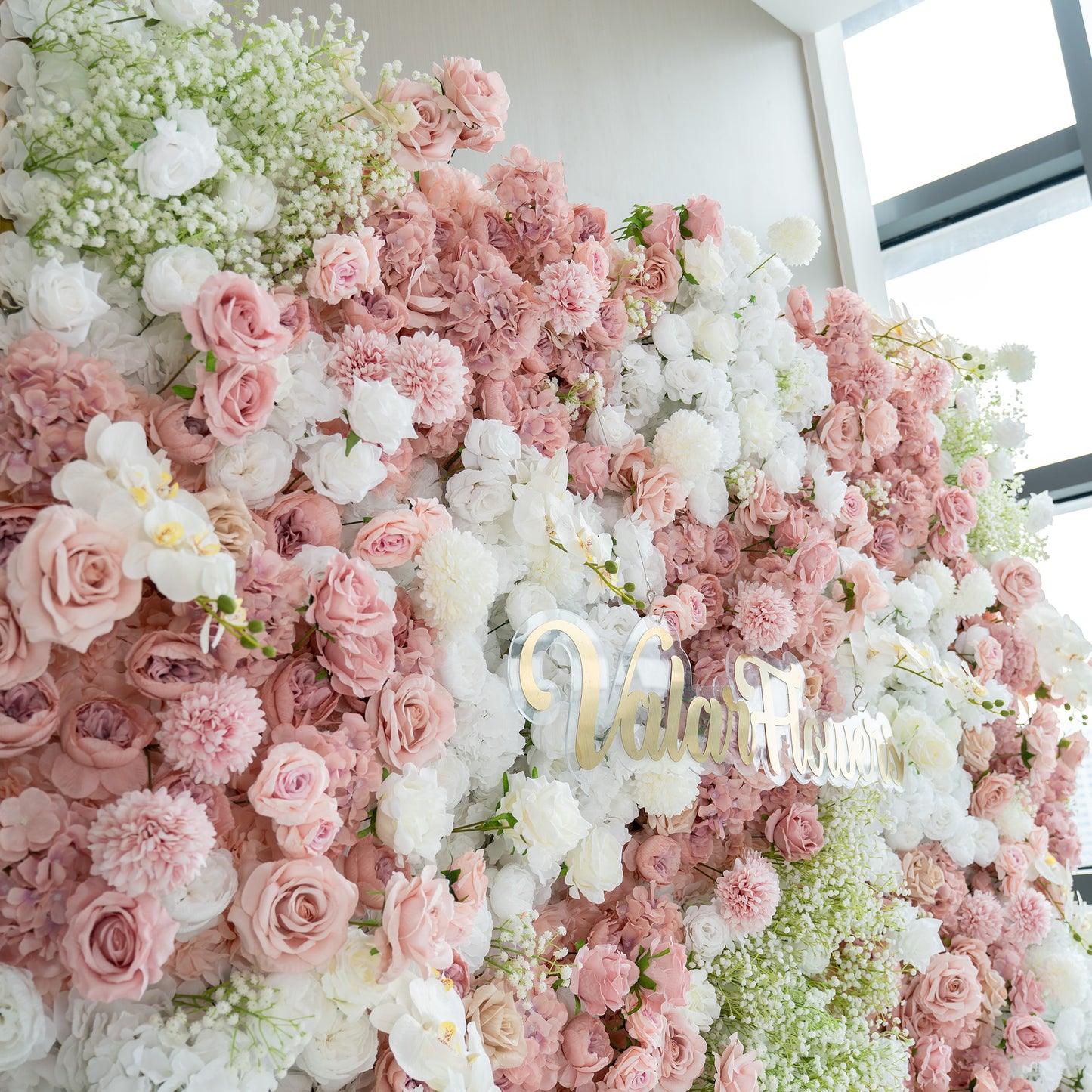 This backdrop is a lush floral arrangement featuring a mix of artificial flowers. It prominently includes roses in various shades of pink and white, along with white orchids and clusters of baby's breath. The color palette is soft and pastel - toned, with gentle pinks and whites. The style is romantic and elegant, ideal for wedding or event decor. The "ValarFlowers" sign adds a branded touch. 