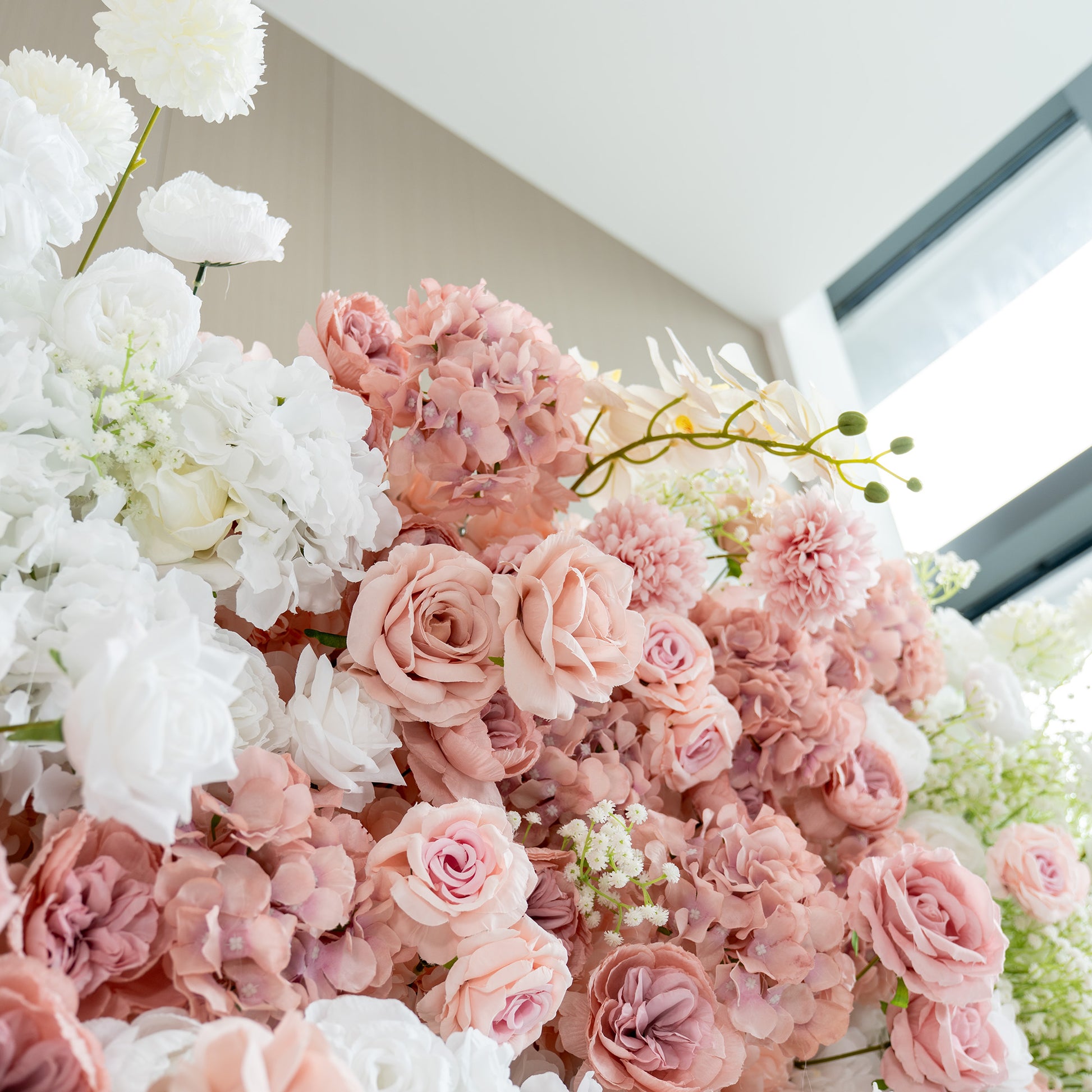 This backdrop features a lush arrangement of artificial flowers. Prominent among them are white and pink roses, white hydrangeas, and delicate sprigs of baby's breath. The color palette is a soft blend of pastel pinks and pure whites, creating a romantic and elegant style, ideal for wedding or bridal events. 