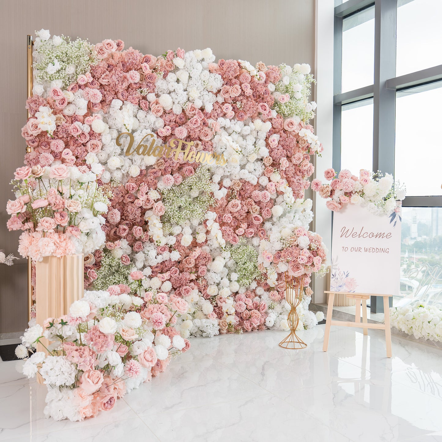 This backdrop is a lush floral arrangement, ideal for a wedding as indicated by the “Welcome to our wedding” sign. It features a mix of artificial roses in shades of pink and white, white chrysanthemums, and clusters of baby's breath. The color palette is soft and pastel - toned, creating a romantic and elegant style. 