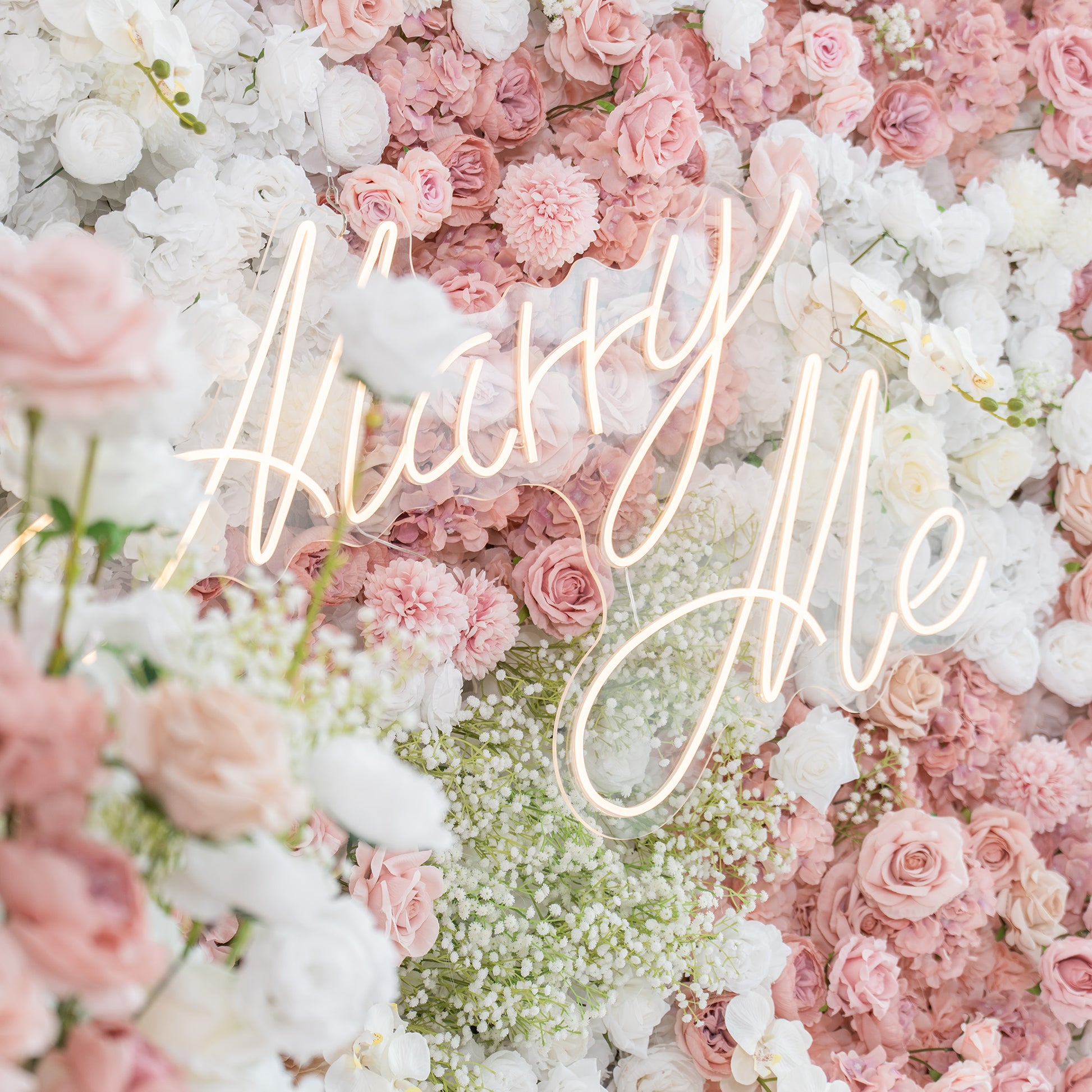 This backdrop is a lush floral wall with a mix of artificial flowers. It includes roses in various shades of pink and white, white chrysanthemums, and clusters of baby's breath. The color palette features soft pastels, creating a romantic and dreamy style. The "Marry Me" neon sign adds a touch of modern romance. 