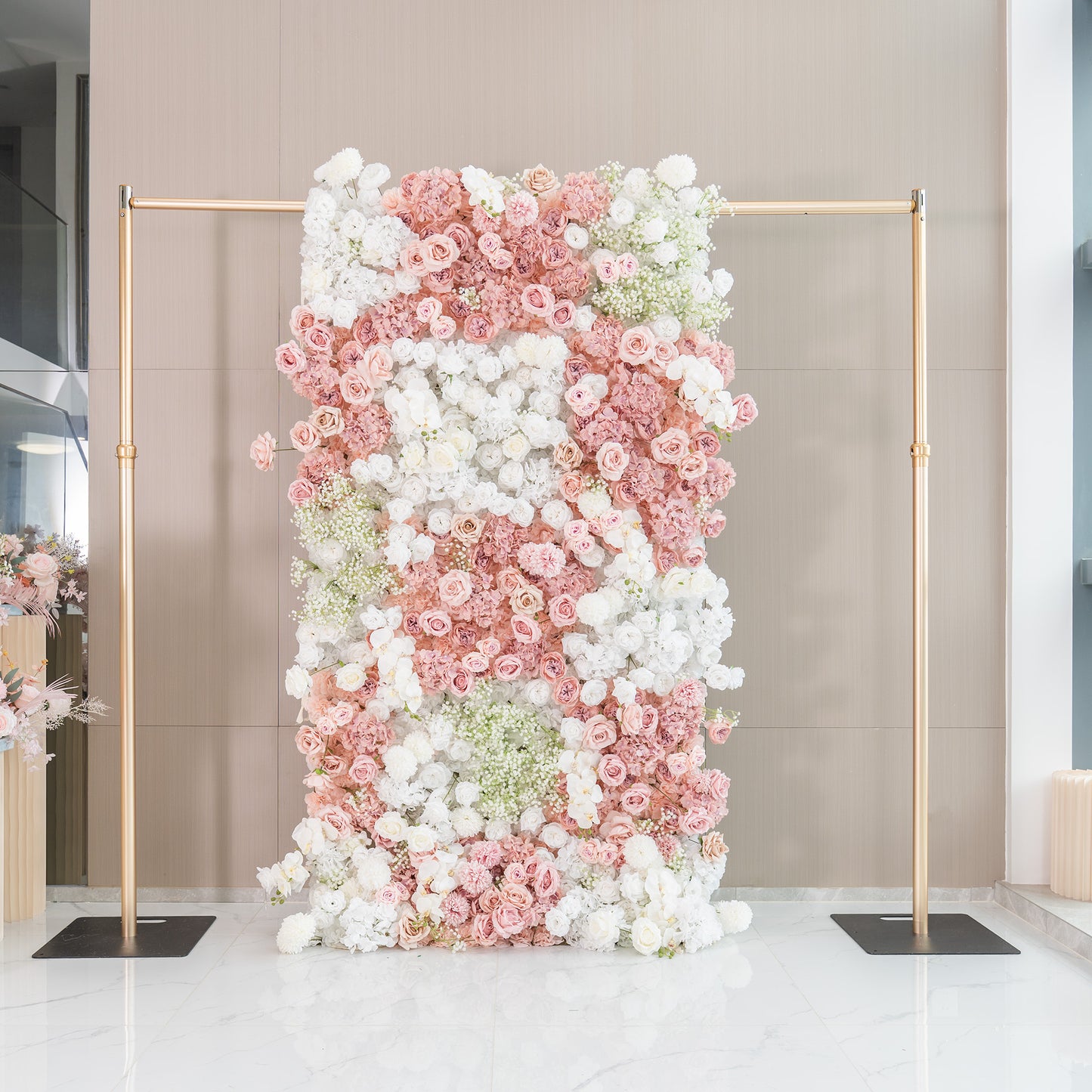 This backdrop is a floral wall supported by golden - hued metal poles. It is densely covered with artificial flowers, including roses in shades of pink and white, white hydrangeas, and clusters of baby's breath. The color palette consists of soft pinks and whites, creating a romantic and elegant style, perfect for wedding or event decor. 