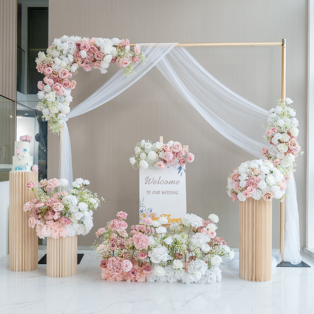This image showcases a charming wedding welcome setup. A golden - framed arch adorned with white fabric drapery serves as the centerpiece. It is decorated with an abundance of artificial flowers in soft pink, white, and hints of green. A sign that reads "Welcome to our wedding" is prominently displayed. Flower arrangements in tall, pleated vases and on the ground enhance the romantic and elegant atmosphere.