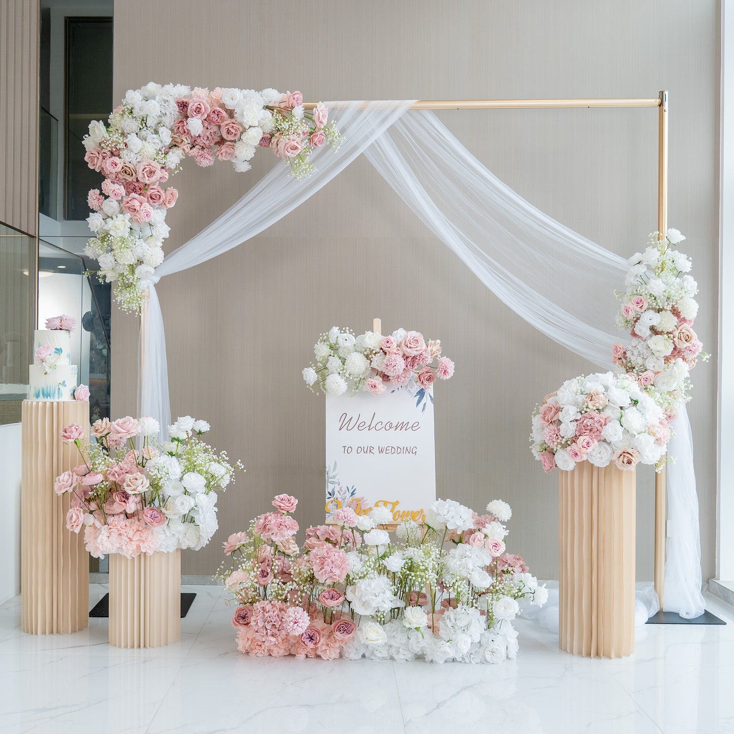 This image showcases a charming wedding welcome setup. A golden - framed arch adorned with white fabric drapery serves as the centerpiece. It is decorated with an abundance of artificial flowers in soft pink, white, and hints of green. A sign that reads "Welcome to our wedding" is prominently displayed. Flower arrangements in tall, pleated vases and on the ground enhance the romantic and elegant atmosphere. 