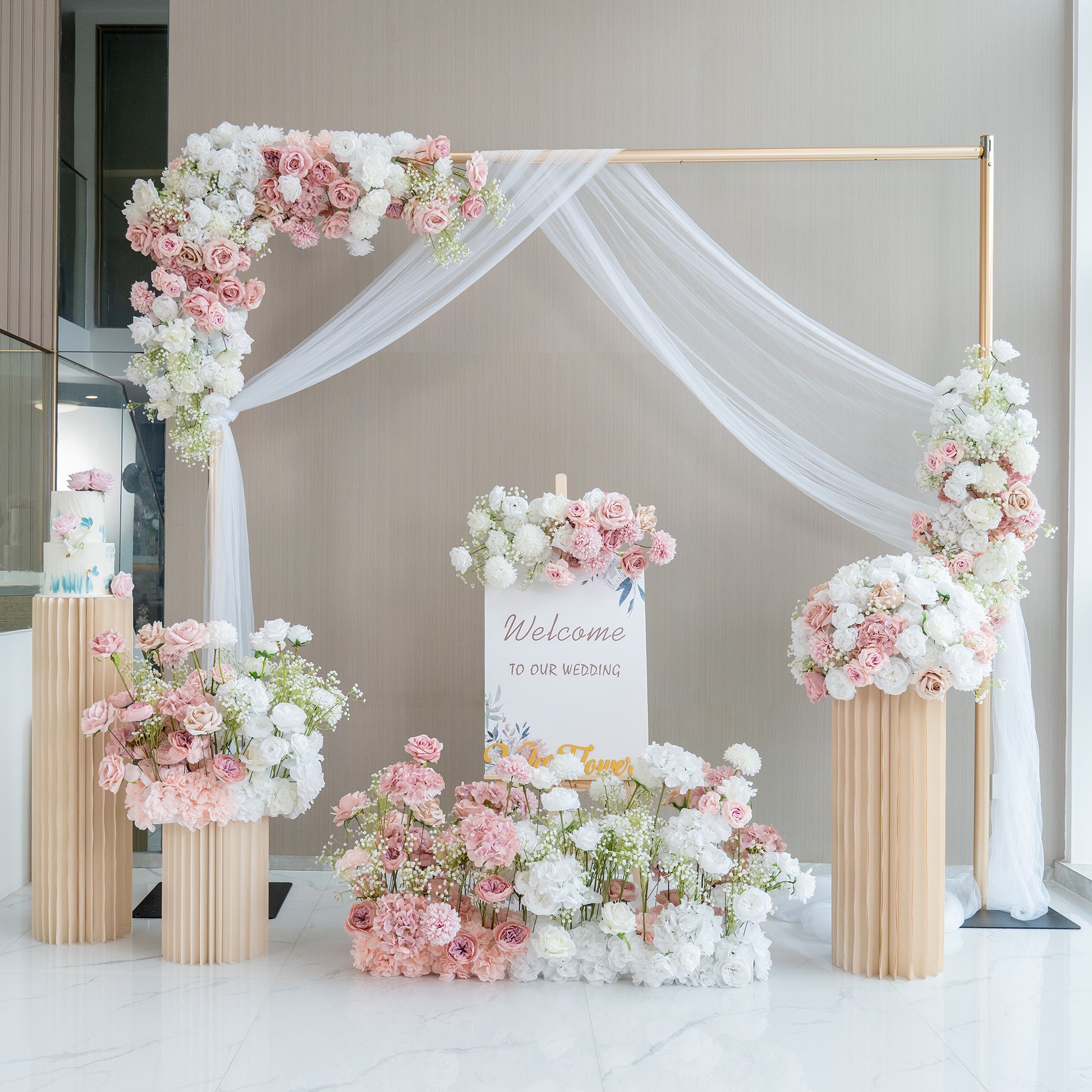 This image showcases a charming wedding welcome setup. A golden - framed arch adorned with white fabric drapery serves as the centerpiece. It is decorated with an abundance of artificial flowers in soft pink, white, and hints of green. A sign that reads "Welcome to our wedding" is prominently displayed. Flower arrangements in tall, pleated vases and on the ground enhance the romantic and elegant atmosphere. 