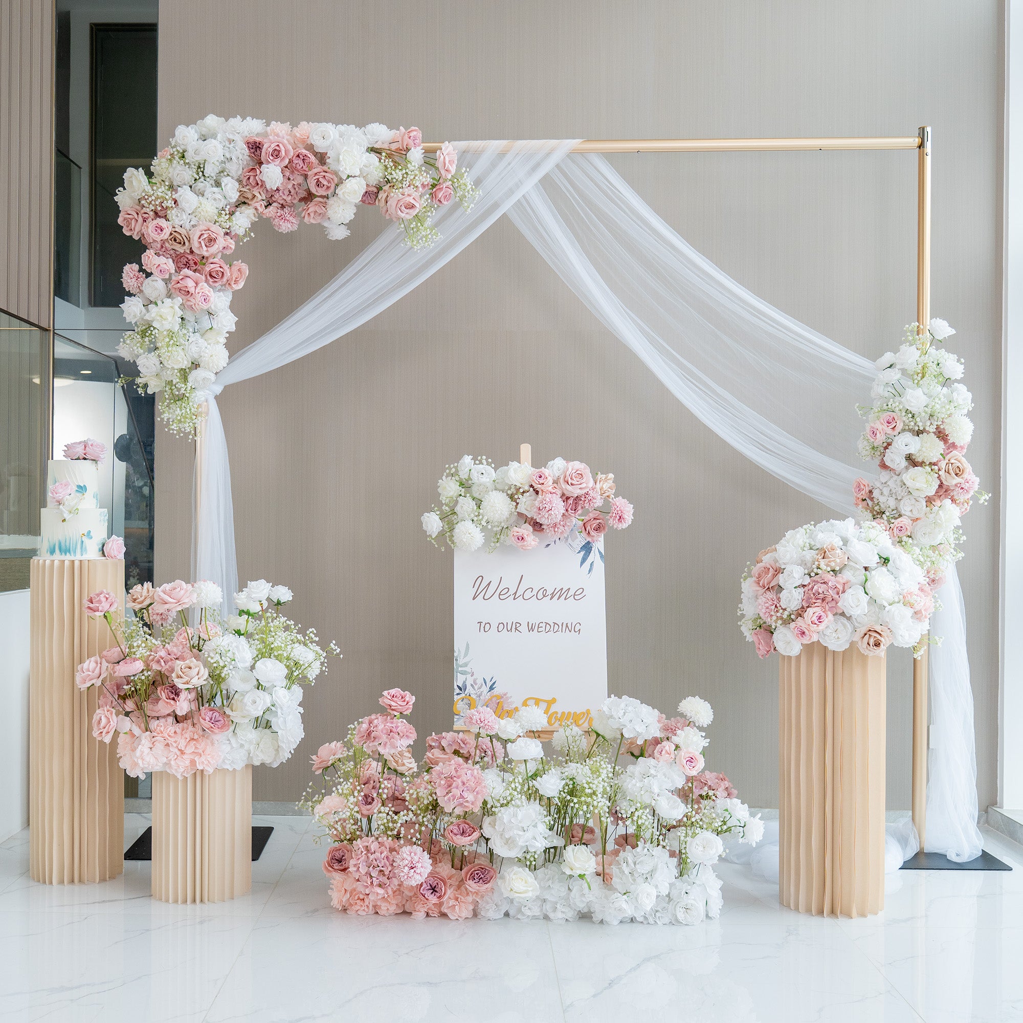 This image showcases a charming wedding welcome setup. A golden - framed arch adorned with white fabric drapery serves as the centerpiece. It is decorated with an abundance of artificial flowers in soft pink, white, and hints of green. A sign that reads &quot;Welcome to our wedding&quot; is prominently displayed. Flower arrangements in tall, pleated vases and on the ground enhance the romantic and elegant atmosphere. 