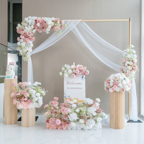 This image showcases a charming wedding welcome setup. A golden - framed arch adorned with white fabric drapery serves as the centerpiece. It is decorated with an abundance of artificial flowers in soft pink, white, and hints of green. A sign that reads "Welcome to our wedding" is prominently displayed. Flower arrangements in tall, pleated vases and on the ground enhance the romantic and elegant atmosphere. 
