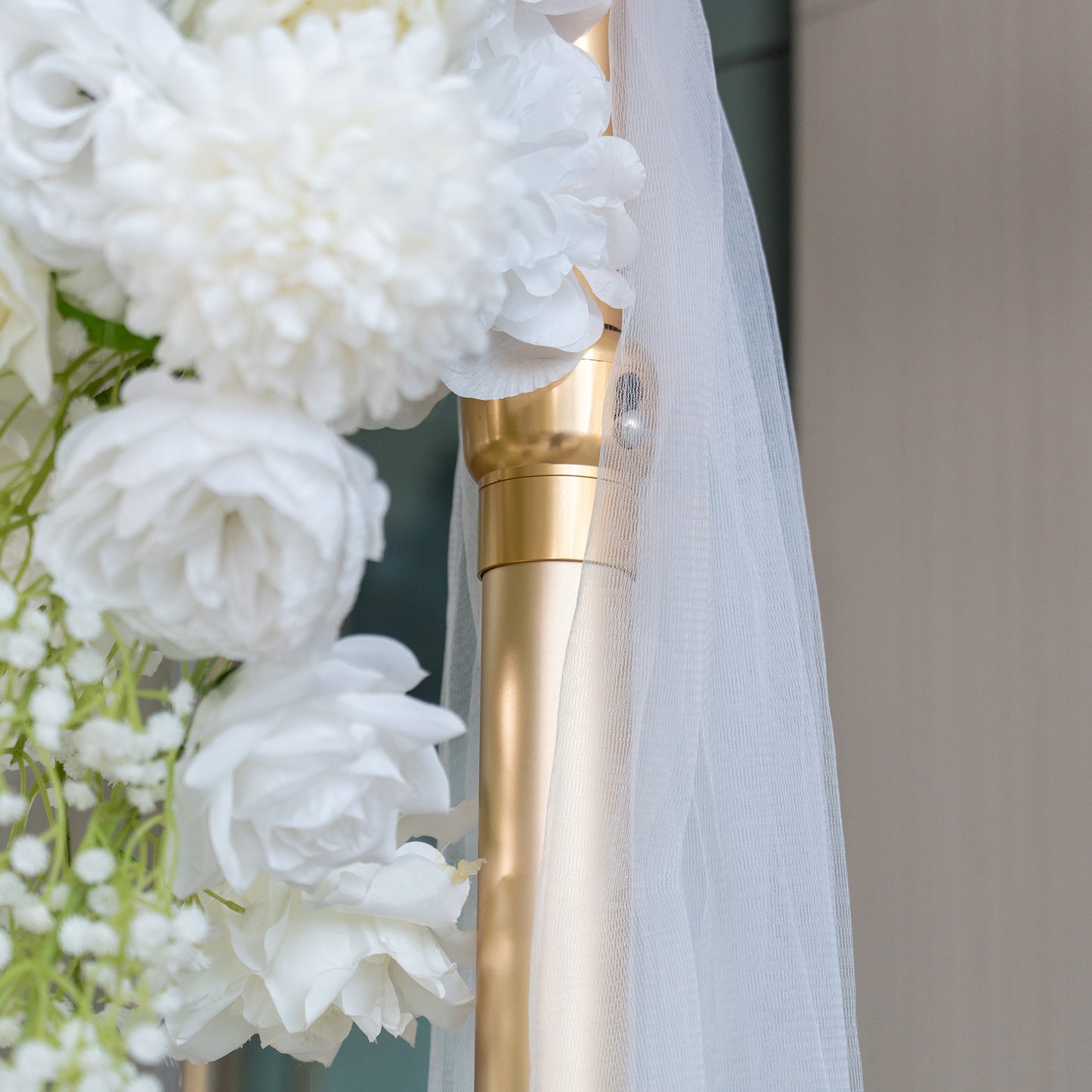This image captures a close - up view of a wedding decor element. A golden metal pole is prominently featured, with a white sheer fabric draped around it. Adjacent to the pole are lush white artificial flowers, including large blooms and delicate baby's breath, creating an elegant and romantic atmosphere typical of wedding arrangements. 