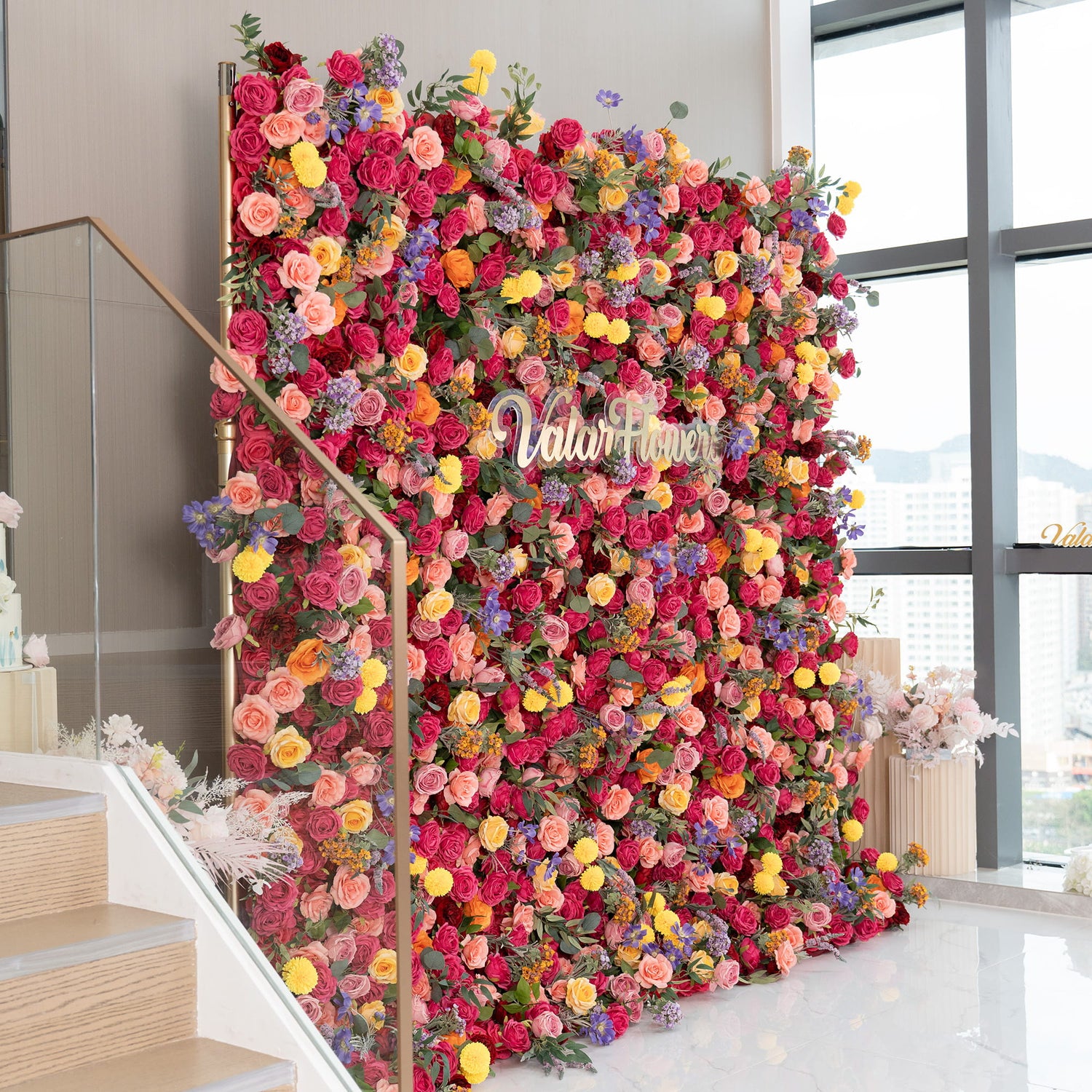 This image features a stunning floral wall adorned with a variety of artificial flowers. Predominant are roses in rich reds, soft pinks, and peaches, complemented by bright yellow pom - pom flowers and delicate purple blooms. The sign &quot;ValarFlowers&quot; stands out. The style is lavish and colorful, perfect for a decorative or event backdrop. 