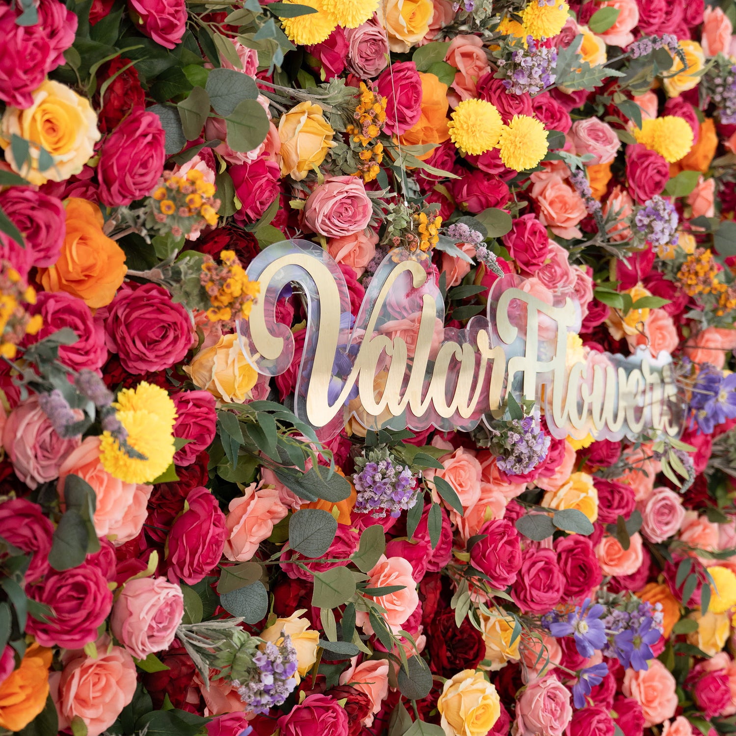 This image showcases a vibrant floral wall densely packed with artificial flowers. Prominent flowers include roses in various shades of red, pink, and peach. There are also bright yellow pom - pom flowers and delicate purple blooms. A sign that reads &quot;ValarFlowers&quot; in elegant, golden - hued lettering stands out. The style is lush and colorful, creating a lively and eye - catching display. 