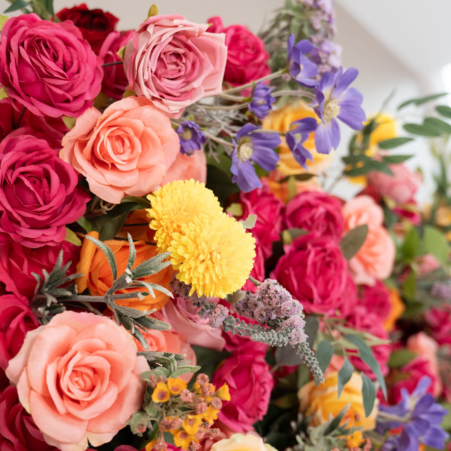This image showcases a vibrant floral arrangement. Prominent flowers include roses in shades of deep red, pink, and peach. There are also bright yellow pom - pom flowers, along with delicate purple and blue blooms. Greenery and small yellow buds add to the mix. The style is colorful and lush, creating a lively and eye - catching display. 