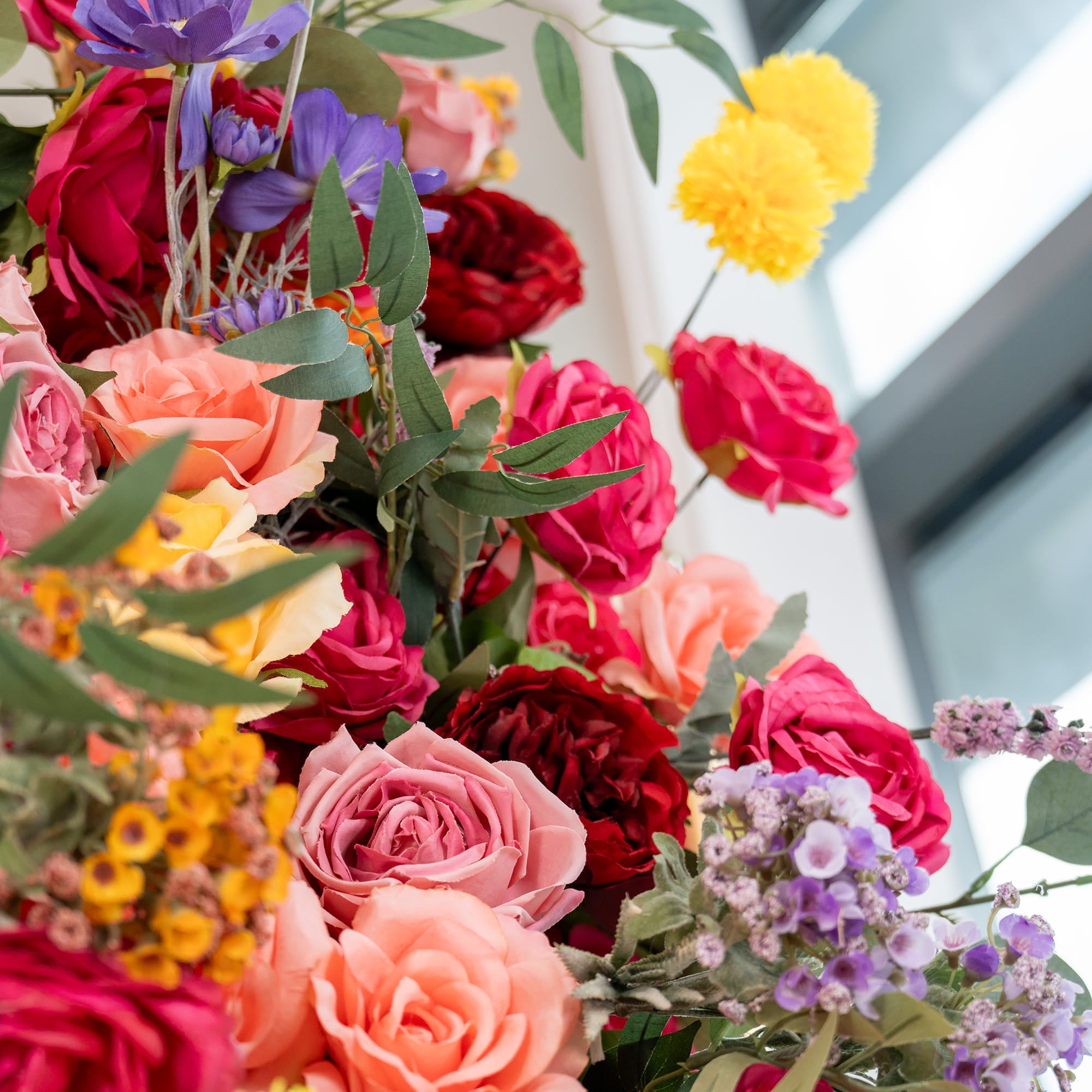 This image features a vibrant floral composition with a variety of artificial flowers. Prominently, there are roses in shades of deep red, pink, and peach. Additionally, bright yellow pom - pom flowers, purple blooms, and small yellow and purple clustered flowers add to the mix. Green foliage complements the colorful display, creating a lush and lively style. 