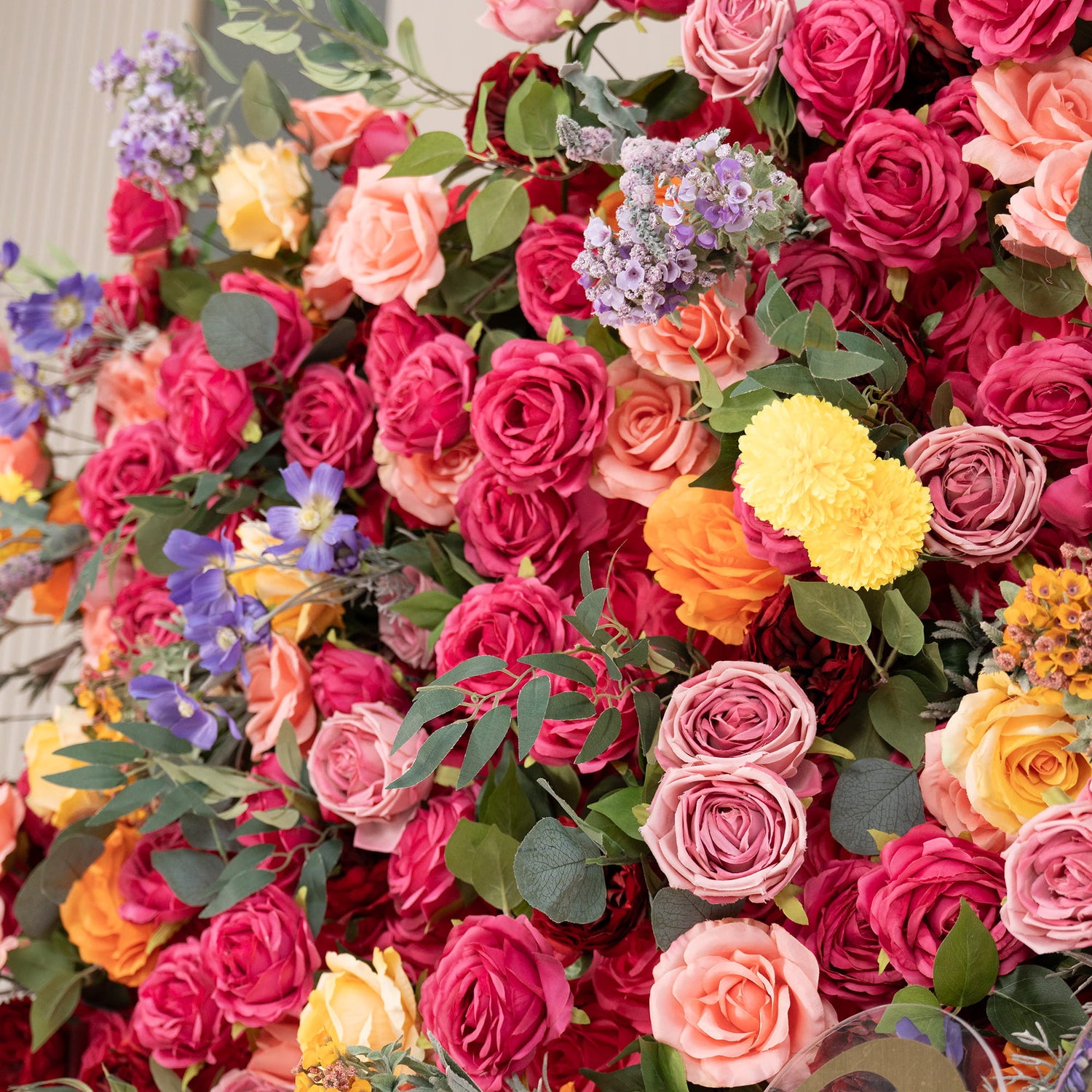 This image showcases a lush floral arrangement. Predominantly, there are roses in a rich palette of colors including deep red, pink, peach, and yellow. Interspersed are vibrant yellow pom - pom flowers, delicate purple blooms, and green foliage. The style is colorful and abundant, creating a lively and eye - catching display. 