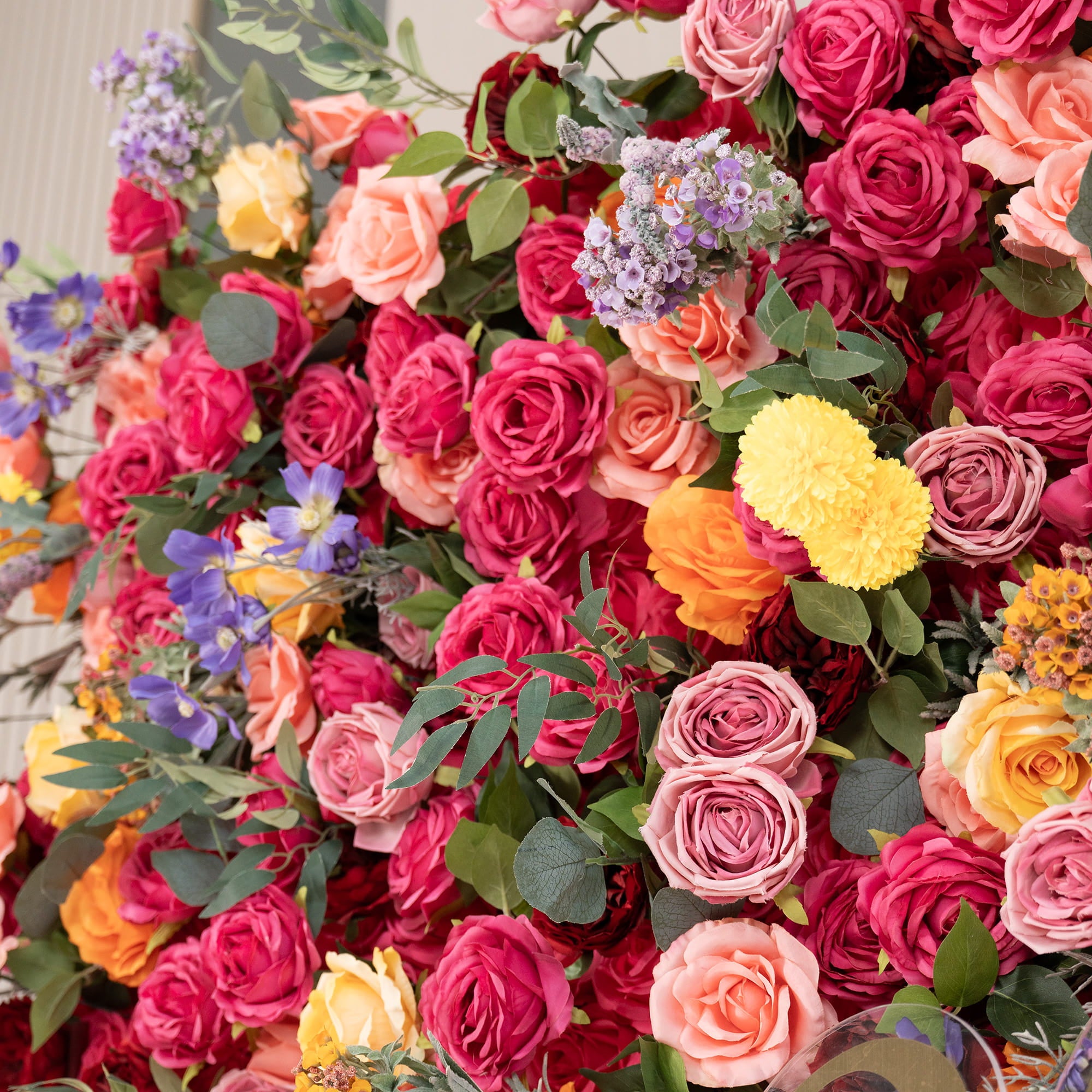 This image showcases a lush floral arrangement. Predominantly, there are roses in a rich palette of colors including deep red, pink, peach, and yellow. Interspersed are vibrant yellow pom - pom flowers, delicate purple blooms, and green foliage. The style is colorful and abundant, creating a lively and eye - catching display. 