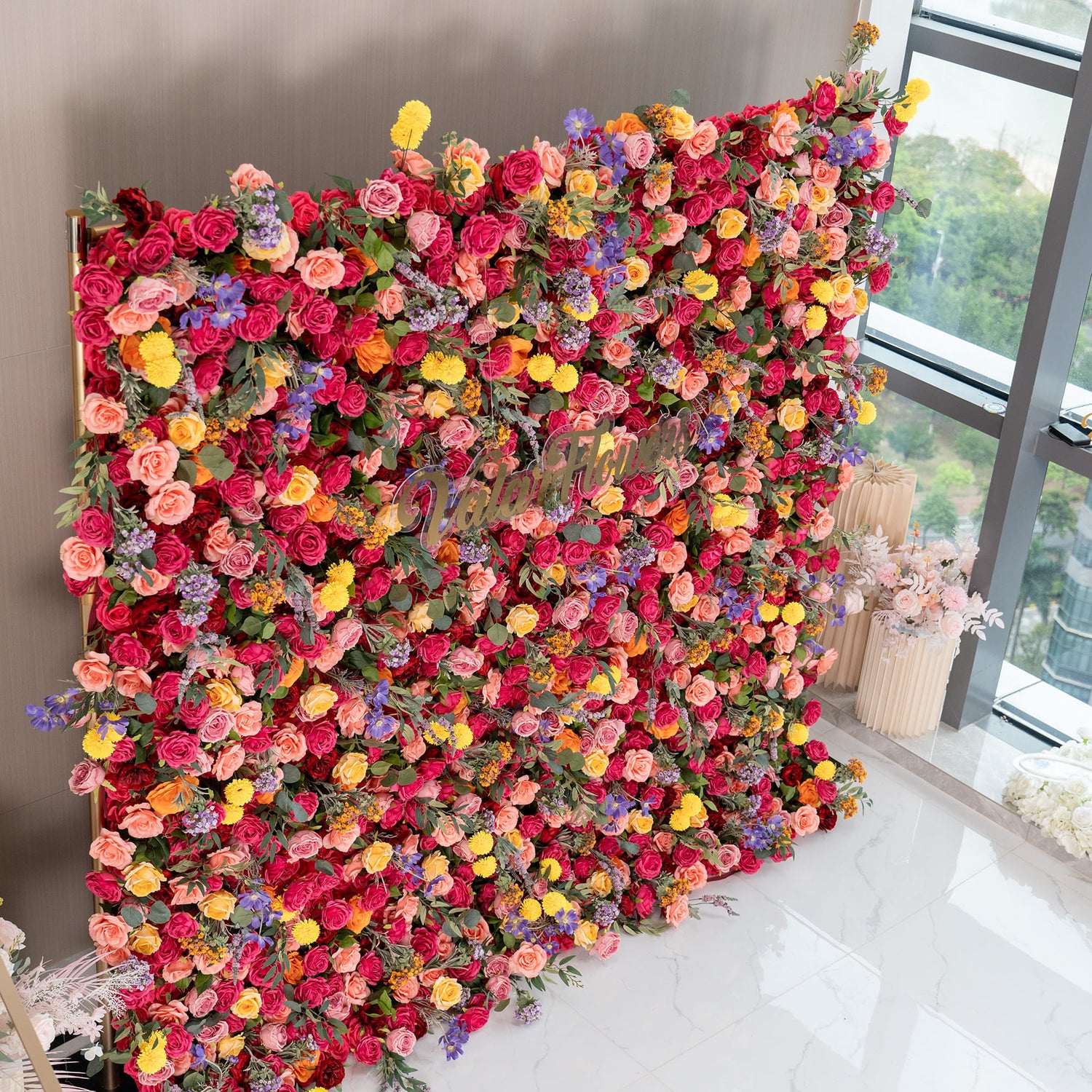 This image features a vibrant floral wall densely packed with artificial flowers. The main flowers are roses in various shades like deep red, pink, and peach. There are also bright yellow pom - pom flowers and delicate purple blooms. The sign &quot;ValarFlowers&quot; is visible. The style is lush and colorful, ideal for event decoration. 