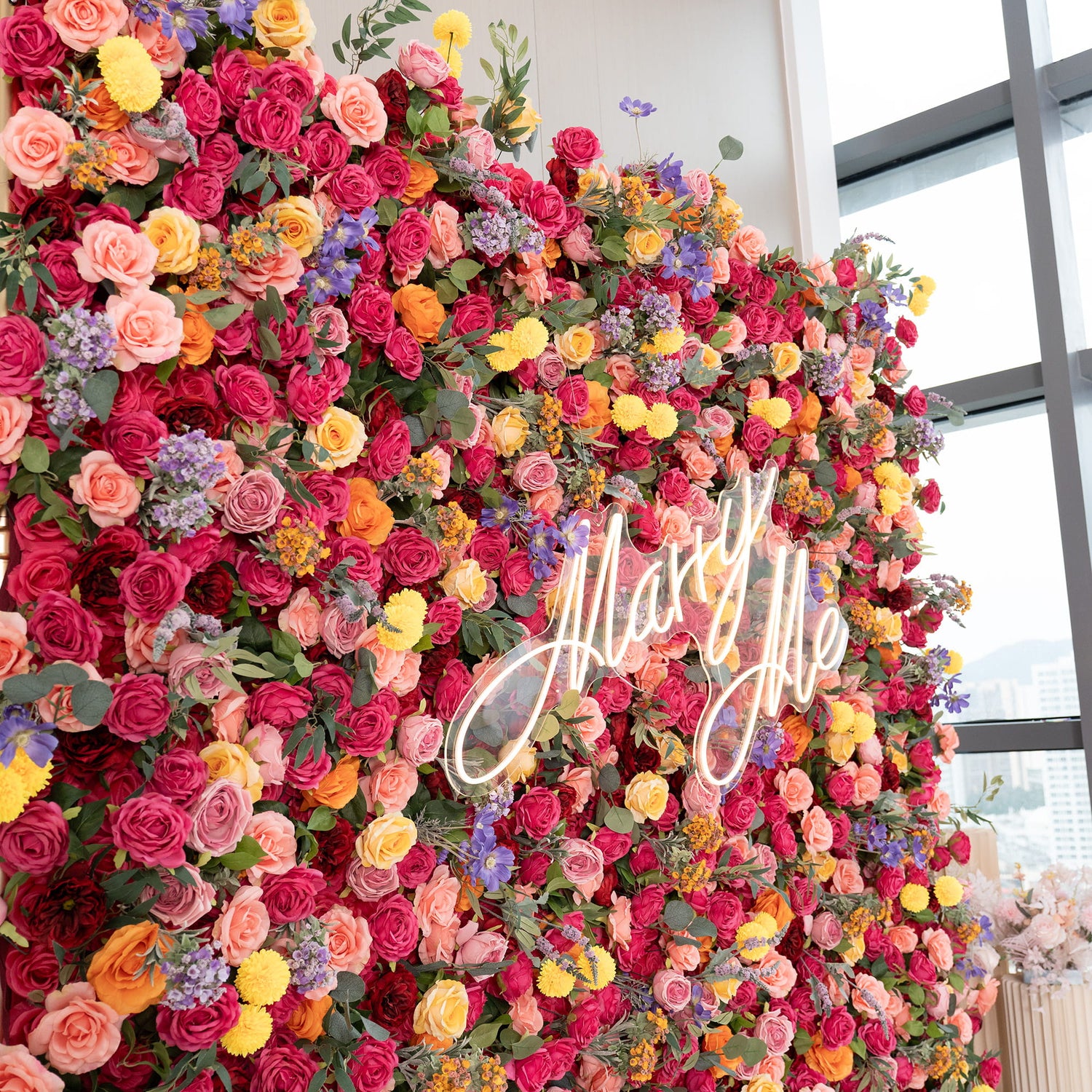 This image showcases a lush floral wall brimming with artificial flowers. Predominantly, there are roses in rich hues of red, pink, peach, and yellow. Interspersed are vibrant yellow pom - pom flowers and delicate purple blooms. A neon sign reading &quot;Marry Me&quot; adds a romantic touch. The style is opulent and colorful, perfect for a special event. 