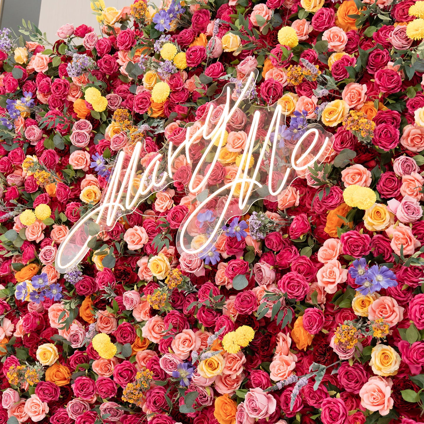 This image showcases a vibrant floral wall densely packed with artificial flowers. Predominant flowers are roses in shades of deep red, pink, peach, and yellow. There are also bright yellow pom - pom flowers and delicate purple and blue blooms. A neon - style sign that reads "Marry Me" stands out. The style is lush, colorful, and romantic. 