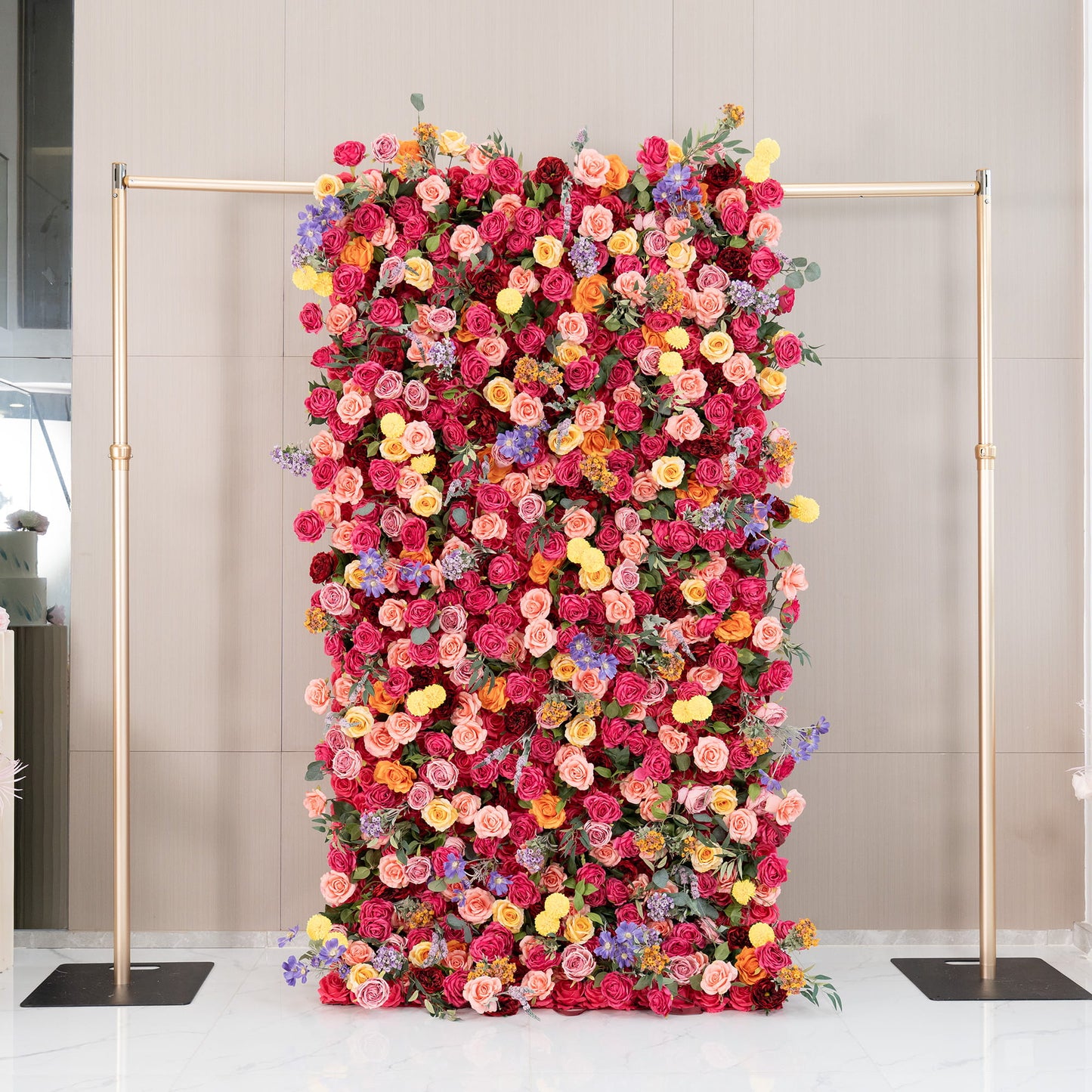 This image features a lush floral wall supported by a metallic gold frame. The wall is densely adorned with artificial flowers, mainly roses in rich shades of red, pink, and peach, along with bright yellow pom - pom flowers and delicate purple blooms. The style is vibrant and romantic, perfect for special events. 