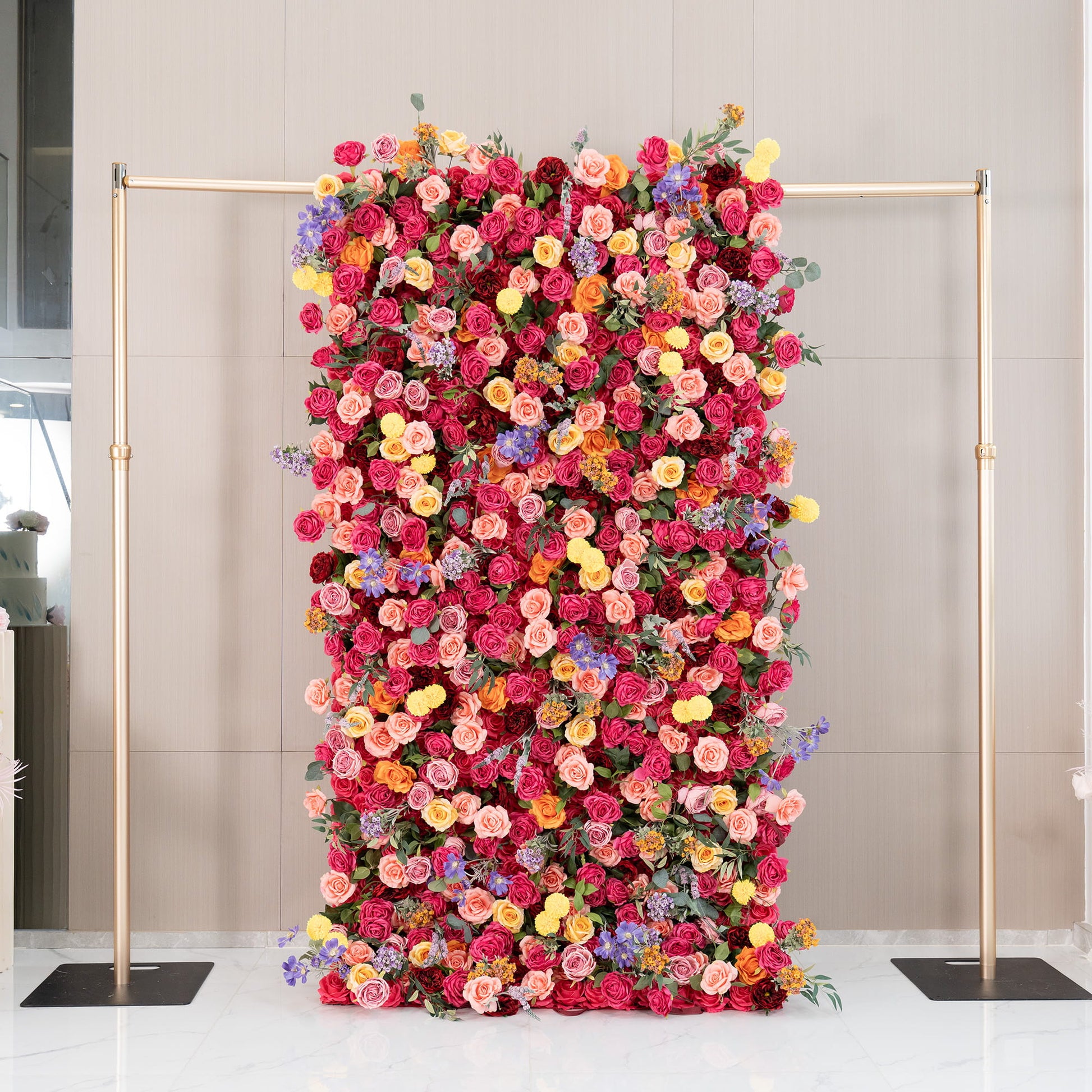 This image features a lush floral wall supported by a metallic gold frame. The wall is densely adorned with artificial flowers, mainly roses in rich shades of red, pink, and peach, along with bright yellow pom - pom flowers and delicate purple blooms. The style is vibrant and romantic, perfect for special events. 