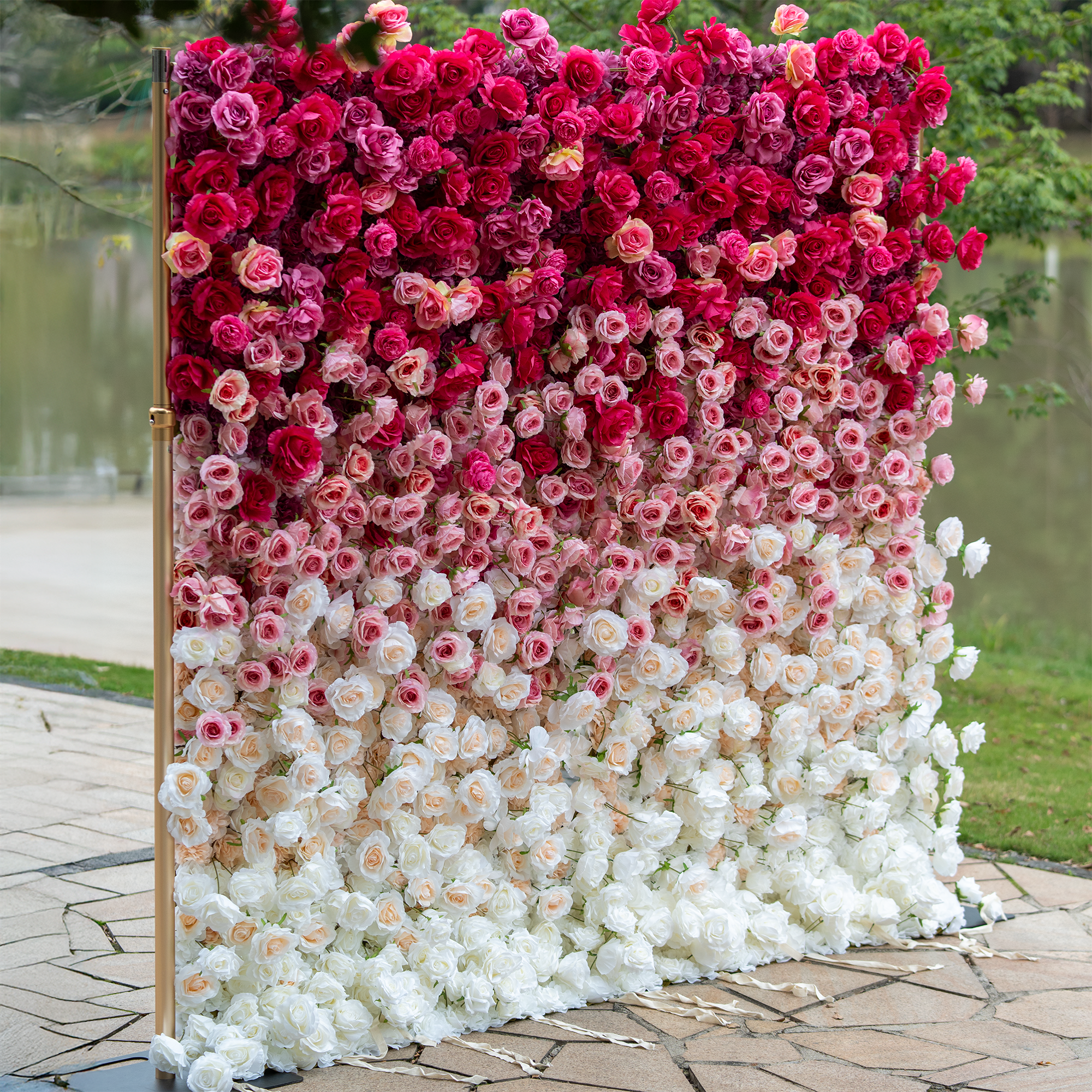 This backdrop is a spectacular display of artificial roses arranged in a gradient from deep red at the top to white at the bottom. Set on a golden stand, it creates a luxurious and romantic scene, ideal for weddings or events, with a serene lakeside and greenery in the background. 