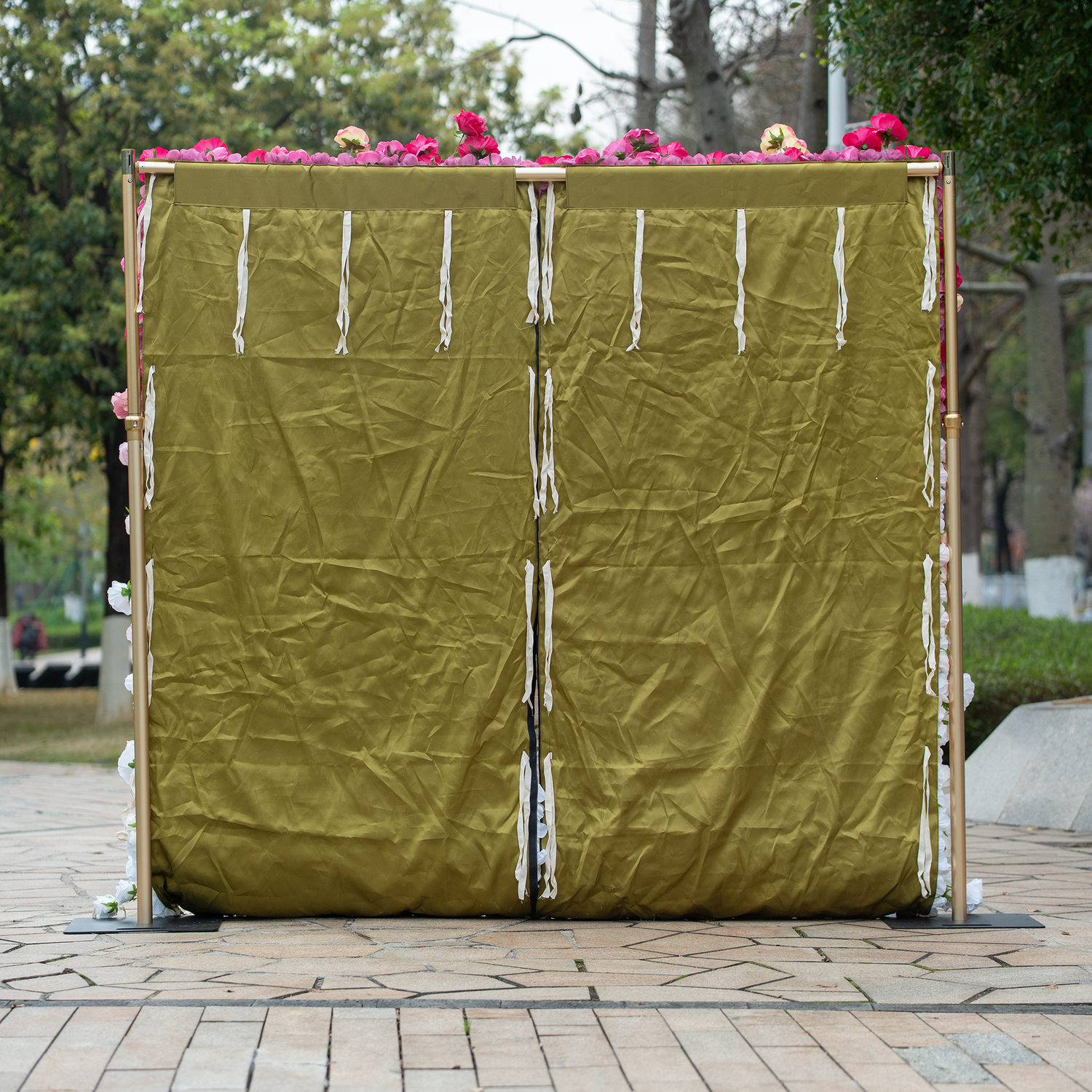 This backdrop features a golden - framed structure with a crumpled, olive - green covering. White, shredded paper strips adorn it, and at the top, there are pink roses. It serves as the backing for a floral display, blending rustic and elegant elements. 