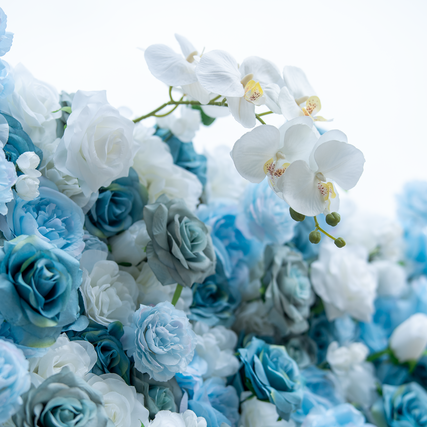 This photo showcases a lush floral arrangement with a harmonious blend of blue and white roses, complemented by elegant white orchids. The soft hues create a serene and sophisticated atmosphere. The dense clustering of flowers highlights their texture and depth, with the orchids' delicate petals and prominent yellow centers adding a touch of refinement. Ideal for upscale events like weddings or galas, it exudes an air of grace and beauty. 