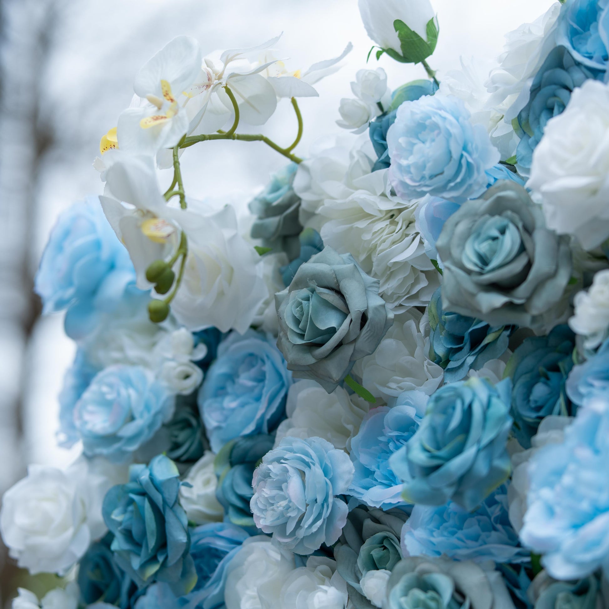 This photo showcases a lush floral arrangement featuring a harmonious blend of blue and white roses, complemented by elegant white orchids. The soft hues create a serene and sophisticated atmosphere. The dense clustering of flowers highlights their texture and depth, with the orchids' delicate petals and prominent yellow centers adding a touch of refinement. Ideal for upscale events like weddings or galas, it exudes an air of grace and beauty. 