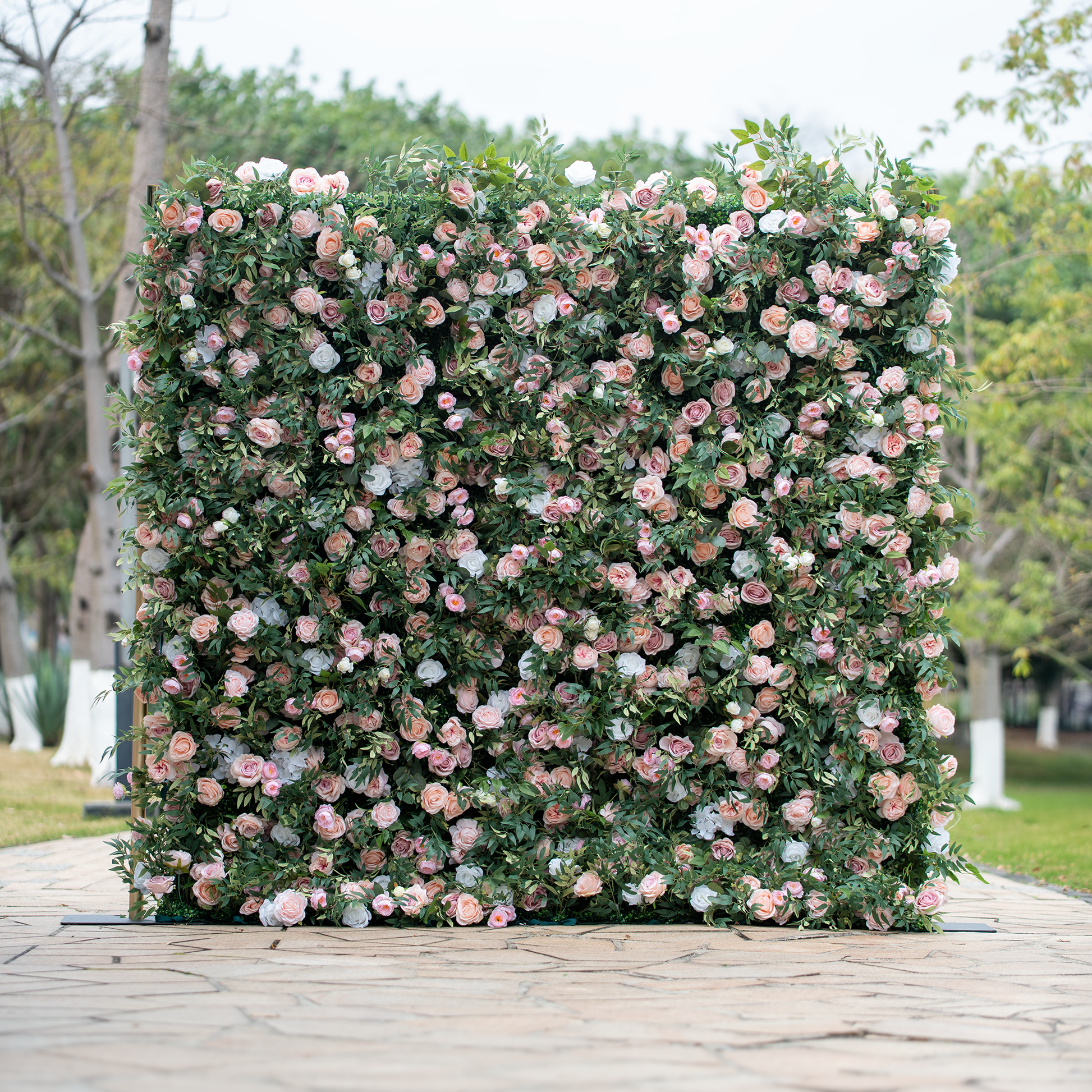 This photo showcases a large, lush floral backdrop densely adorned with an array of pink and white roses interspersed among abundant green foliage. The flowers are tightly clustered, creating a rich, textured appearance. Set outdoors on a paved area with trees in the background, it exudes a romantic and elegant ambiance, making it an ideal decorative element for weddings, garden parties, or other celebratory events. 