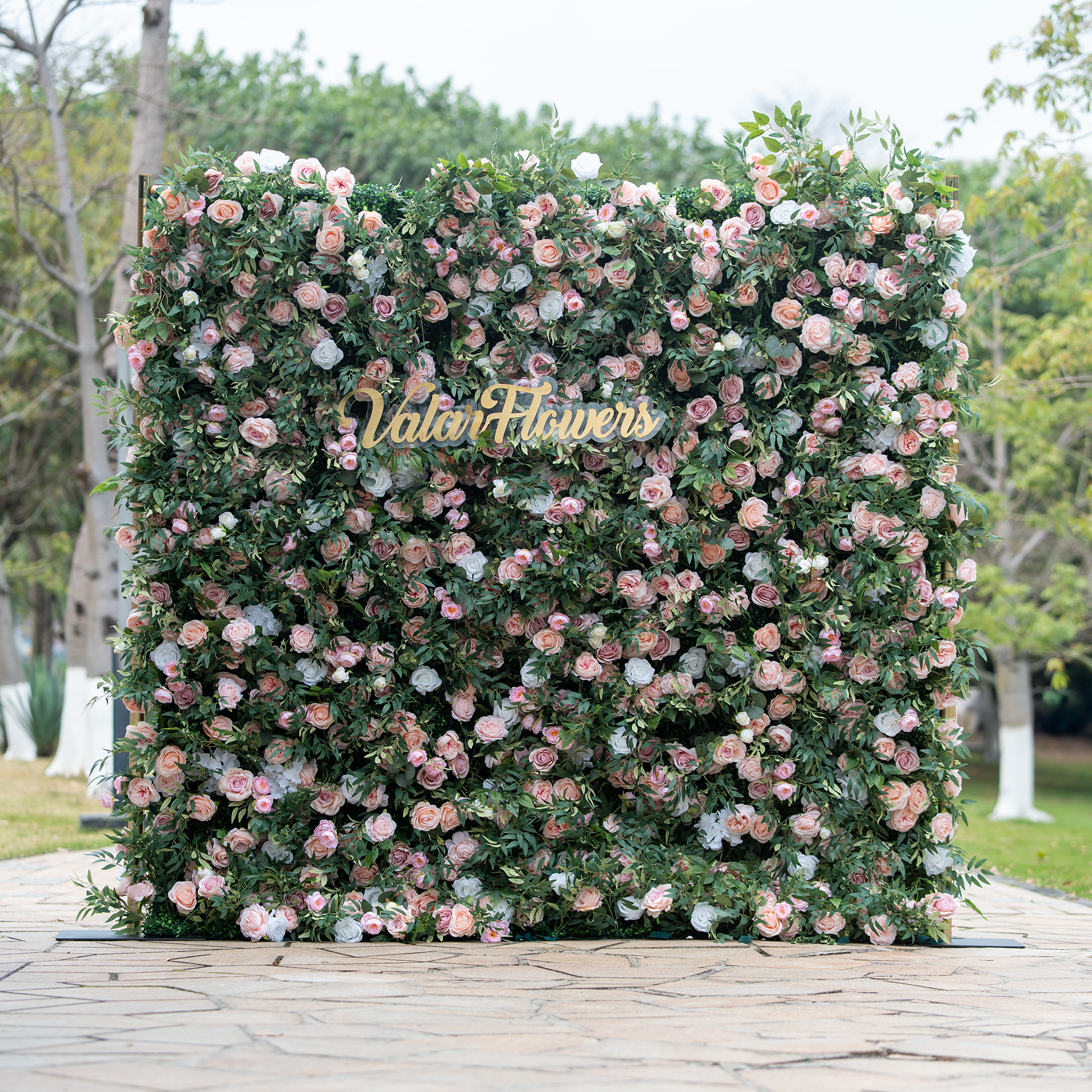 This photo showcases a large, lush floral backdrop densely adorned with pink and white roses interspersed among green foliage. The flowers create a rich, textured appearance. In the center, golden text reads "ValarFlowers," adding a touch of branding. Set outdoors with trees in the background, the backdrop exudes elegance, making it perfect for events like weddings or upscale garden parties. 