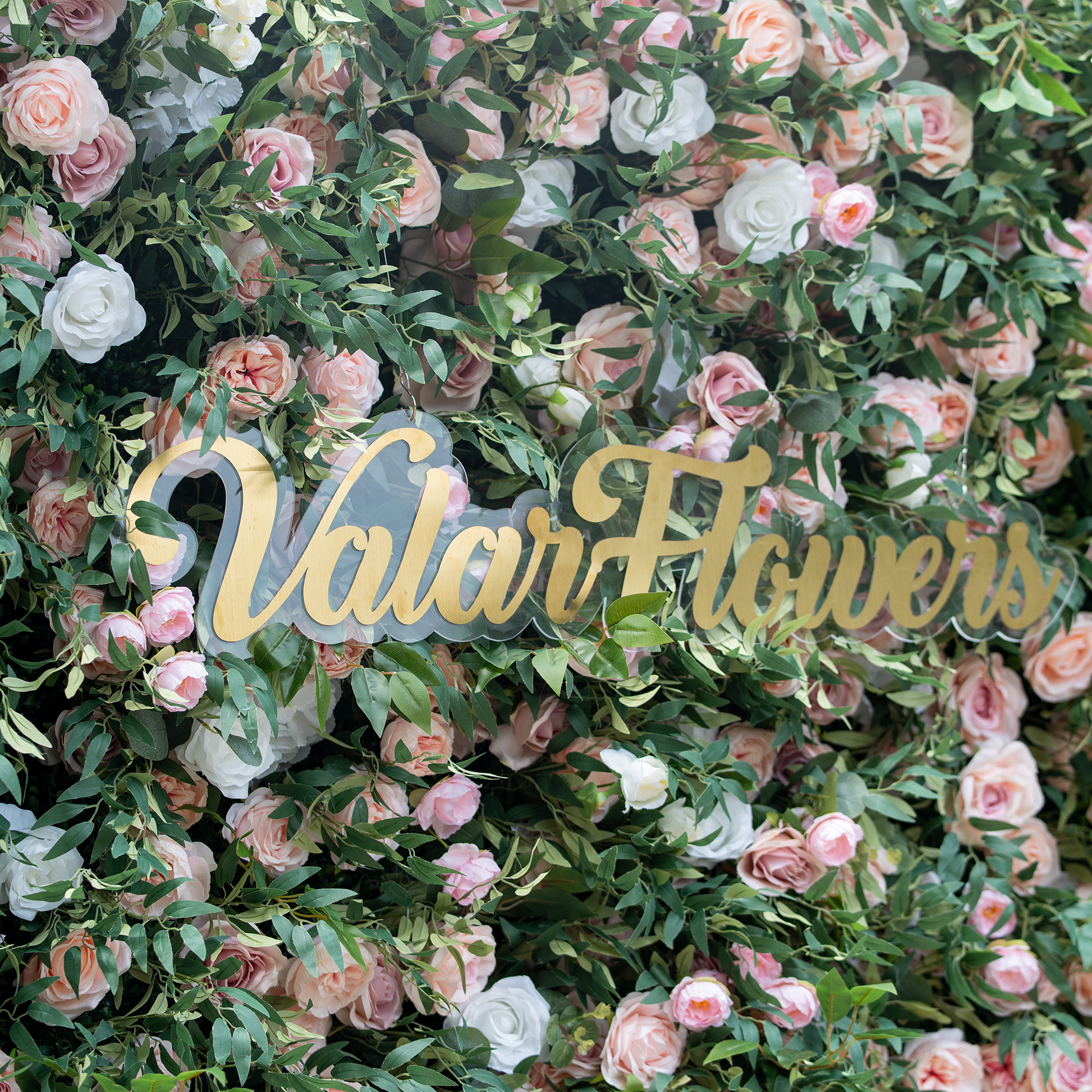This photo showcases a dense floral backdrop composed of pink and white roses interspersed with lush green foliage. Prominently displayed in the center is the text "ValarFlowers" in elegant golden lettering. Ideal for events like wedding receptions or a "Romantic Garden Festival", the soft - hued roses and verdant leaves create a dreamy, sophisticated atmosphere, perfect for celebratory and refined occasions. 