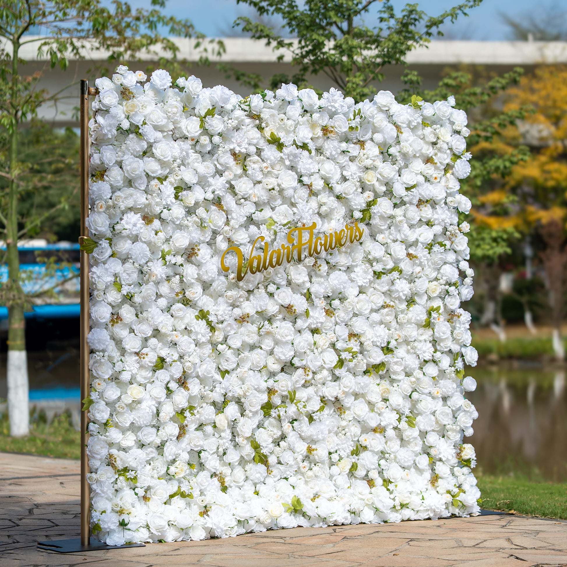 This floral backdrop is densely adorned with artificial white roses, interspersed with small green leaves. The uniform white color of the flowers creates a clean, elegant, and sophisticated look. The golden “ValarFlowers” sign adds a touch of luxury. The overall style is classic and pure, suitable for formal events such as weddings or upscale parties. 
