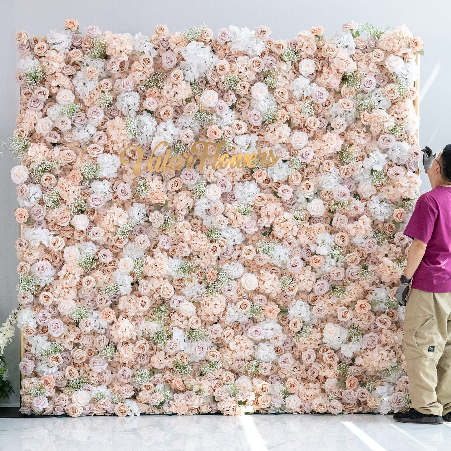 This floral backdrop is a lush, full - coverage arrangement. It consists mainly of artificial roses in soft hues like blush, cream, and light pink, interspersed with white hydrangeas and delicate baby's breath - like greenery. The color palette is gentle and pastel, creating a romantic and elegant style, ideal for events such as weddings or photo booths. The “ValarFlowers” sign adds a branded touch. 