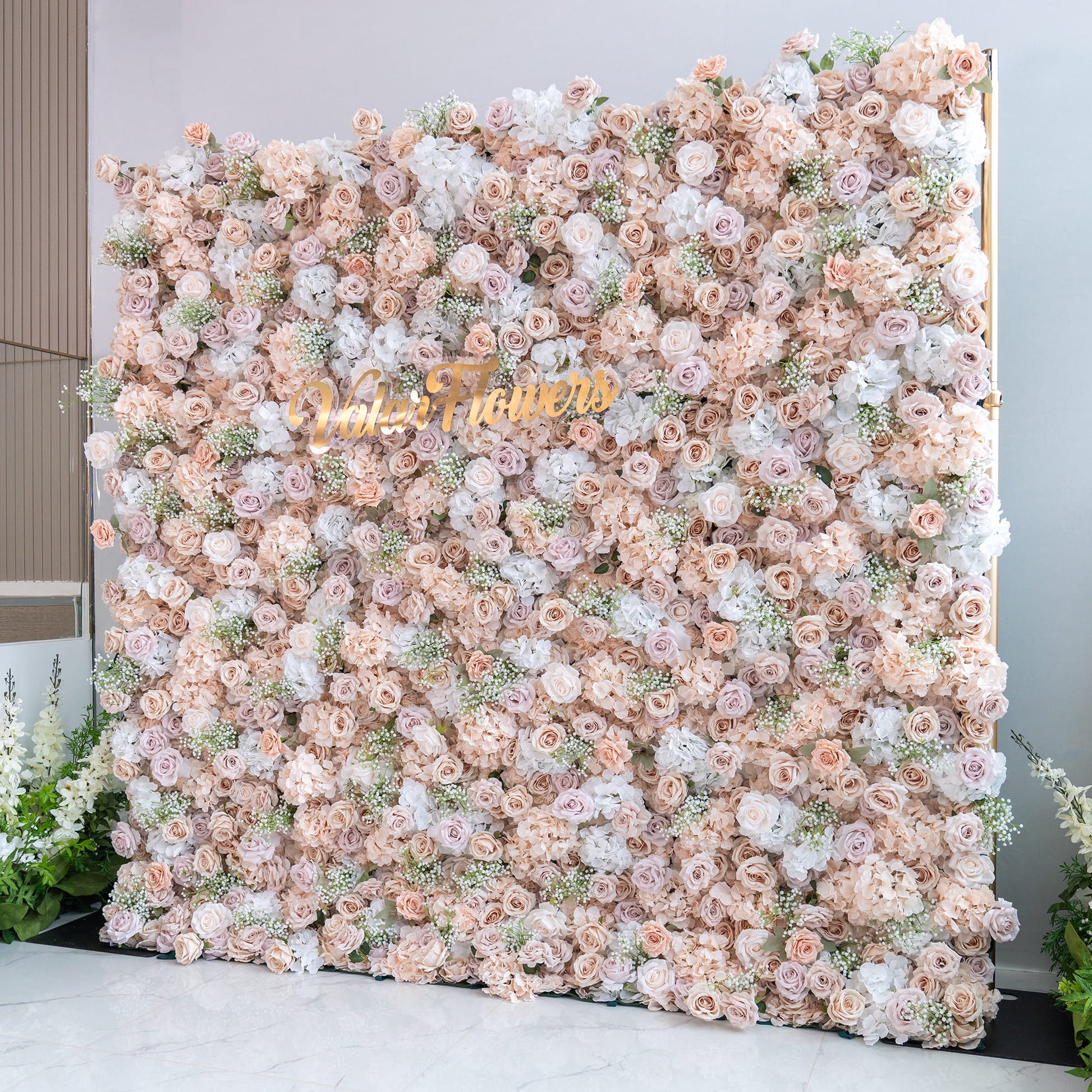 This backdrop is a lush floral wall filled with artificial flowers. It features roses in various shades of blush, cream, and pale pink, interspersed with white hydrangeas and delicate sprigs of baby's - breath - like greenery. The color palette is soft and warm, creating a romantic and elegant style, perfect for events like weddings or photo shoots. The “ValarFlowers” sign adds a branding element. 