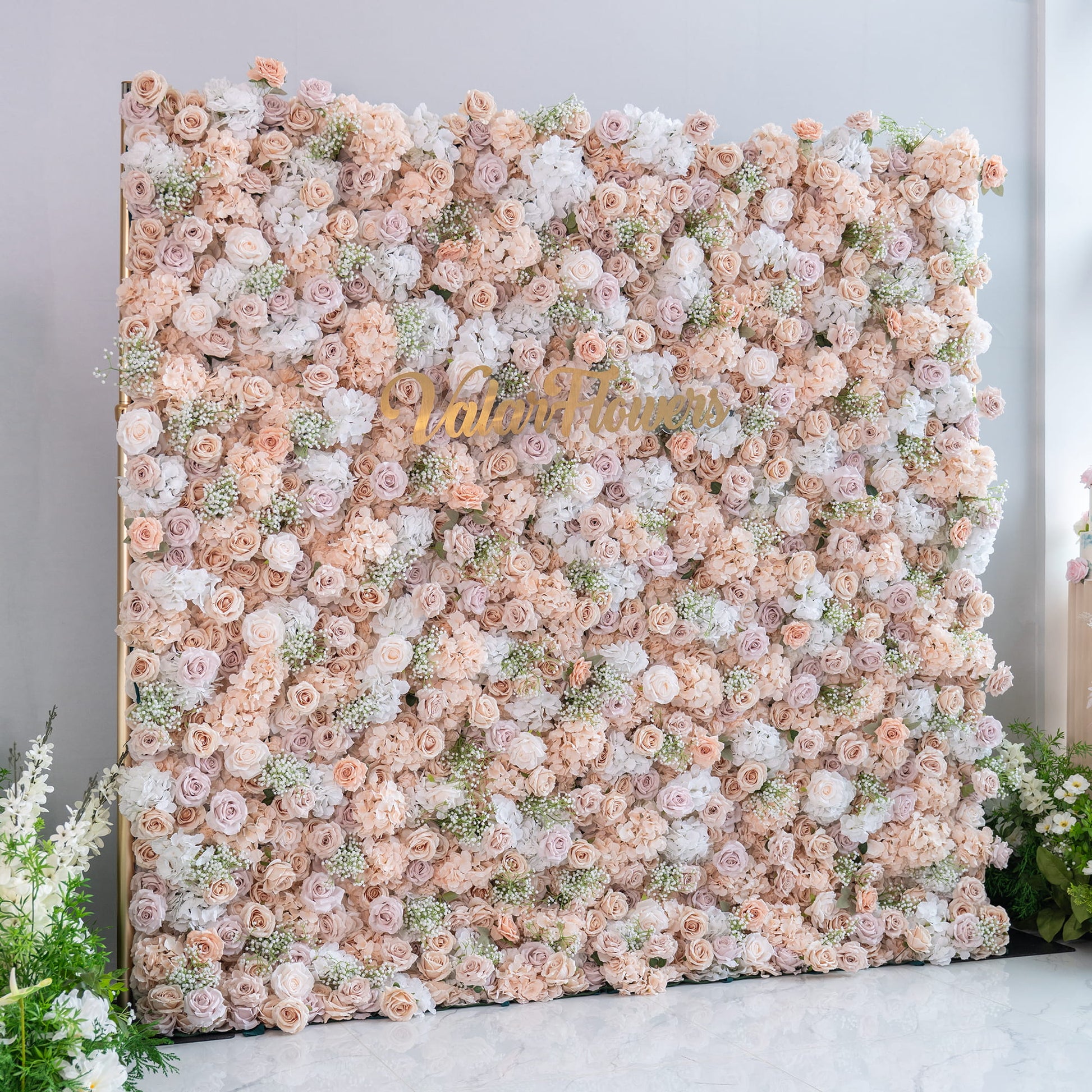 This floral backdrop is densely packed with artificial flowers, primarily featuring roses in various shades of blush, cream, and white. Interspersed are small clusters of baby's breath - like greenery. The color palette is soft and neutral, creating an elegant, romantic style, perfect for wedding or event photo backdrops. The "ValarFlowers" sign adds a branding touch. 
