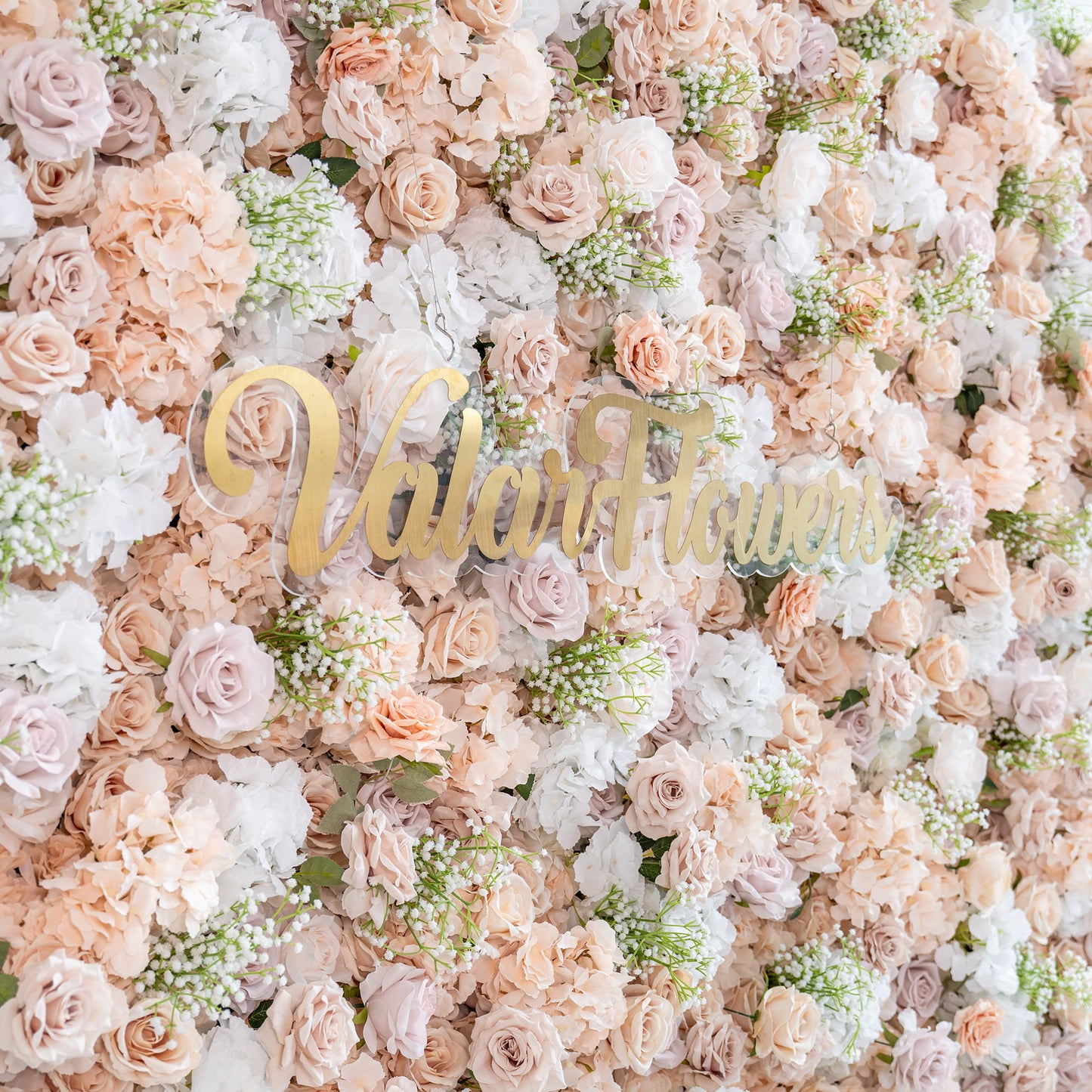 This floral backdrop is a lush, textured display. It features artificial roses in a palette of soft blush, cream, and pale pink hues, interspersed with white hydrangeas and delicate sprigs of baby's - breath - like greenery. The overall style is romantic and elegant, perfect for events such as weddings or bridal showers. The "ValarFlowers" signage adds a touch of branding. 