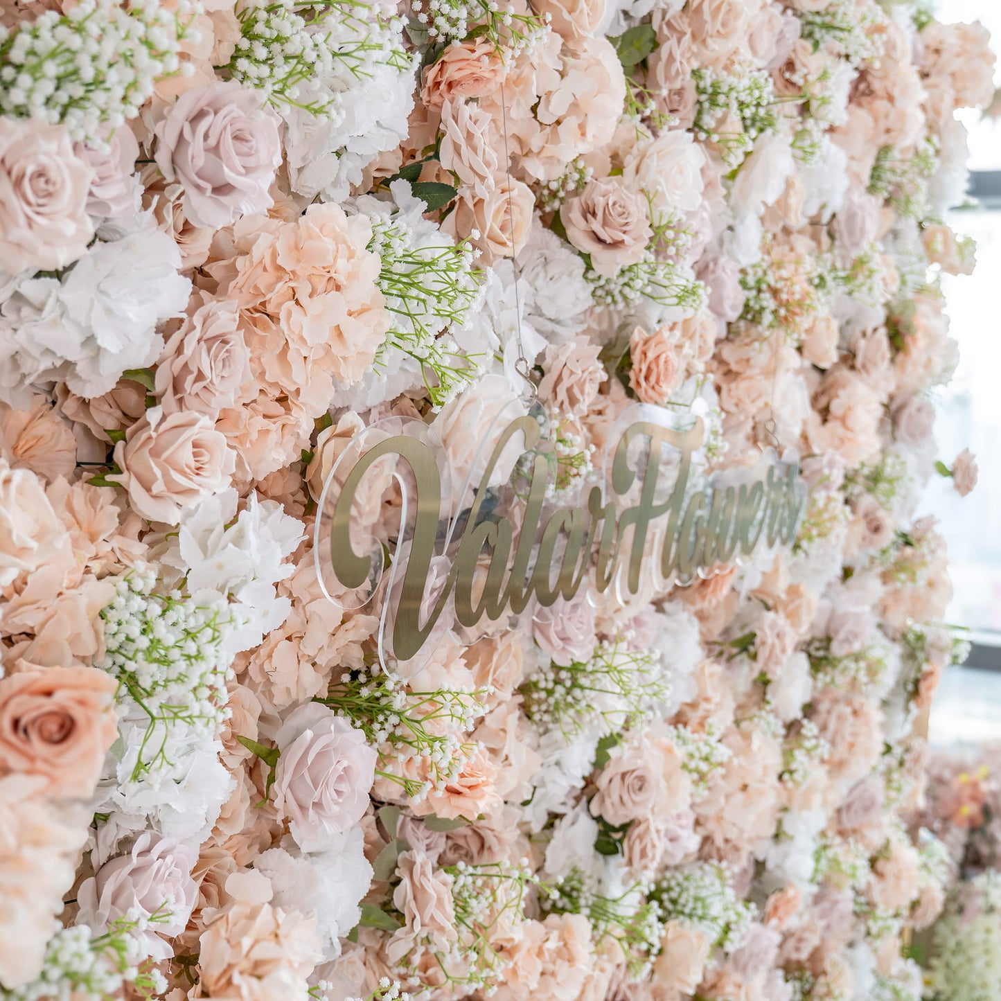 This floral backdrop is a lush composition of artificial flowers. It prominently features roses in various shades of blush, cream, and soft pink, interspersed with white hydrangeas and delicate sprigs of baby's - breath - like greenery. The color palette is gentle and warm, creating a romantic and elegant style, ideal for events like weddings or photo shoots. The "ValarFlowers" sign adds a branded touch. 