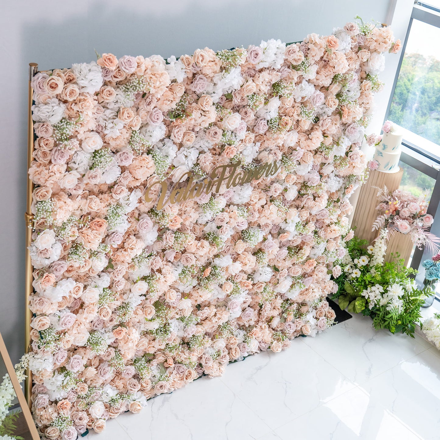 This floral backdrop is a dense arrangement perfect for events. It features artificial roses in soft shades of blush, cream, and pale pink, interspersed with white hydrangeas and delicate sprigs of baby's - breath - like greenery. The color palette is gentle and warm, creating a romantic and elegant style. The “ValarFlowers” sign adds a branded touch. 