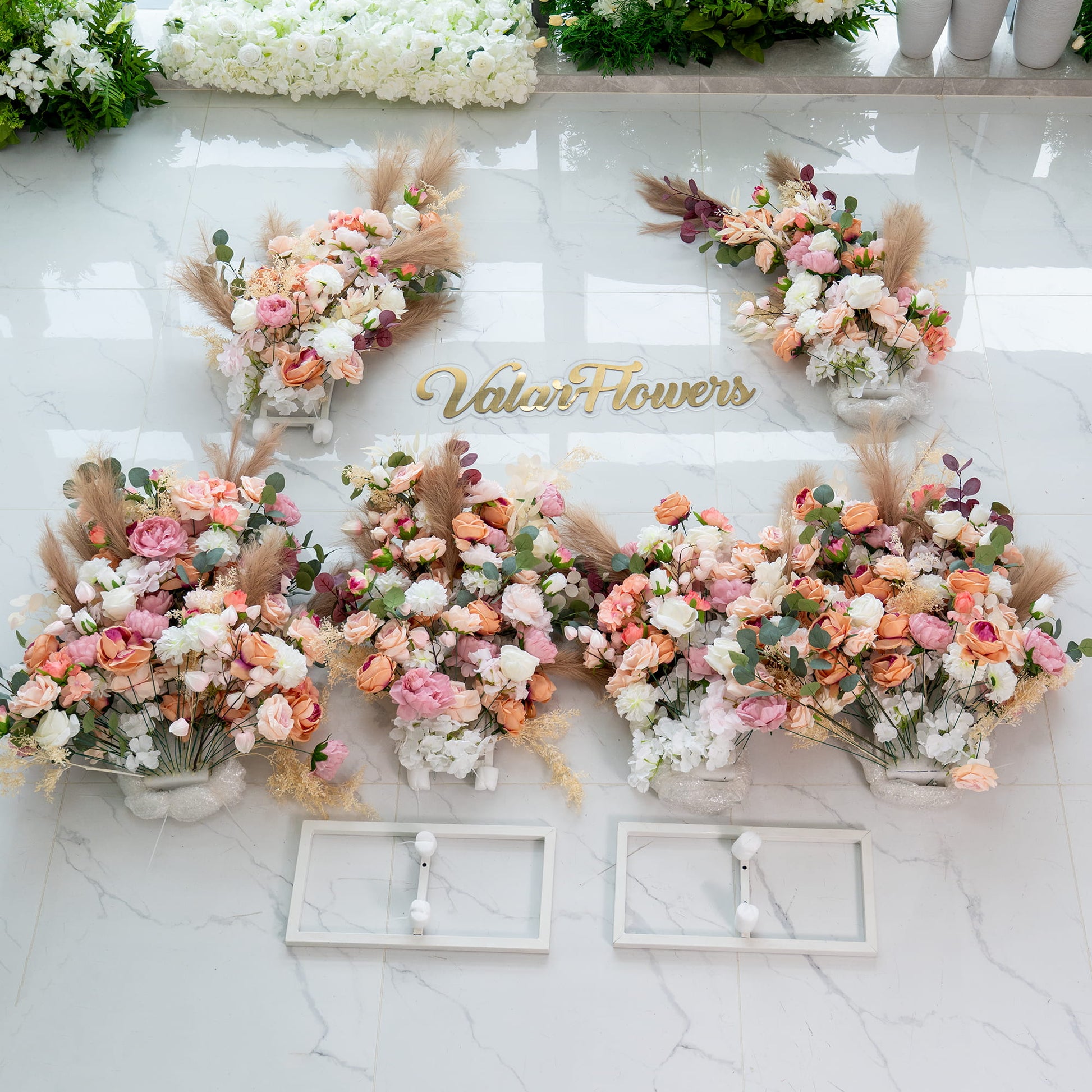 This backdrop features several lush floral arrangements placed on a marble - like floor. The flowers come in a soft palette of pinks, peaches, and whites, with varieties like roses and peonies. Pampas grass and eucalyptus leaves add texture. There are also structural elements and a sign that reads “Valar Flowers”. The overall style is romantic and elegant, perfect for a wedding or event. 