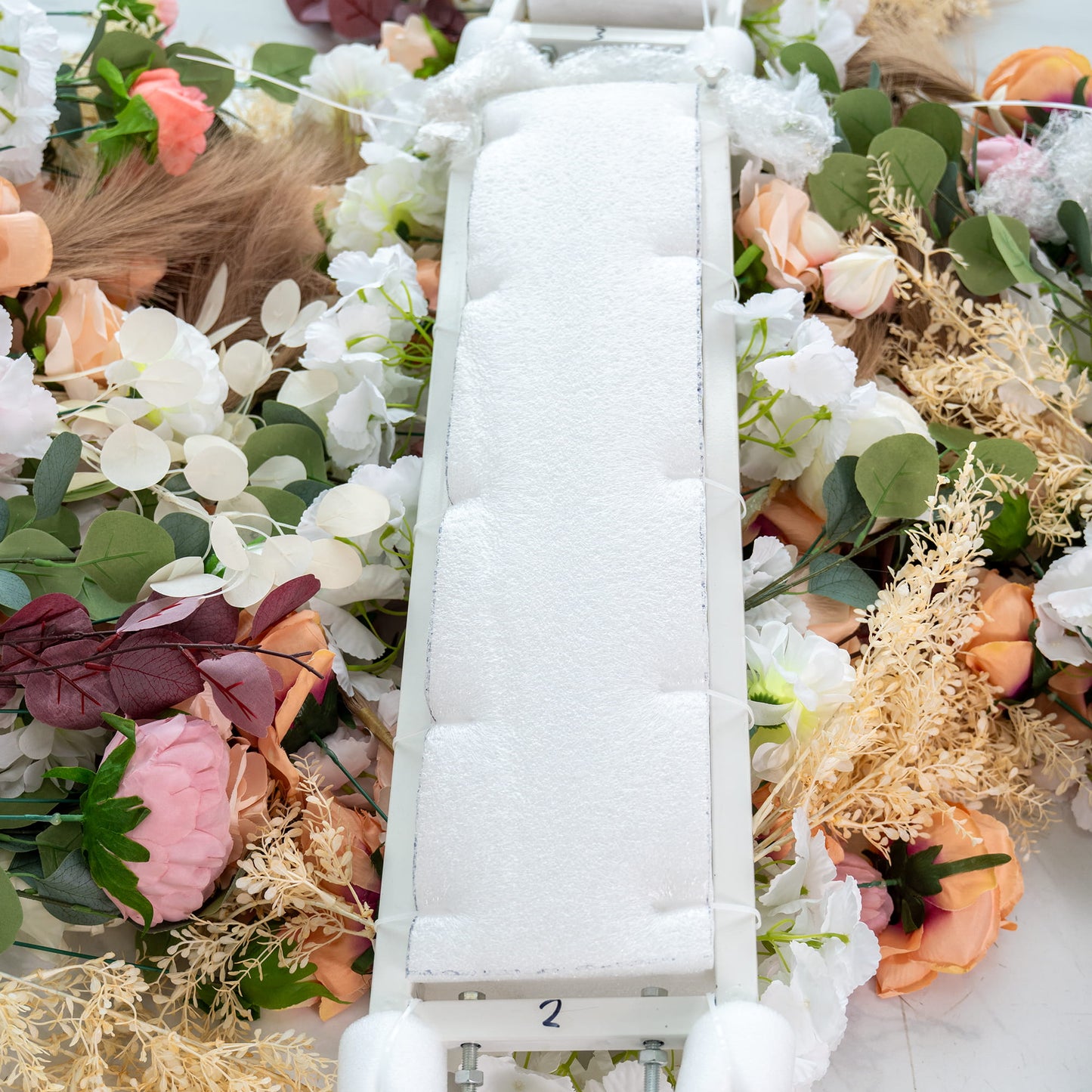 This image shows a white - cushioned structure, numbered "2," surrounded by a variety of artificial flowers in shades of white, pink, peach, and green foliage. The flowers include roses, peonies, and other assorted blooms, along with pampas - like elements. It appears to be part of a floral arrangement setup, possibly for an event such as a wedding. 